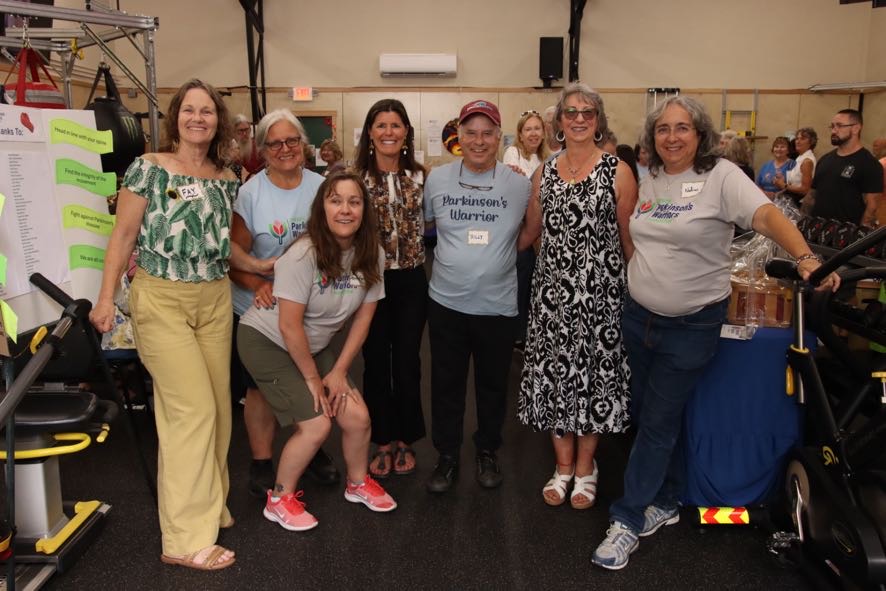 Group of eight women standing together indoors, smiling, with fitness equipment and other people in the background.