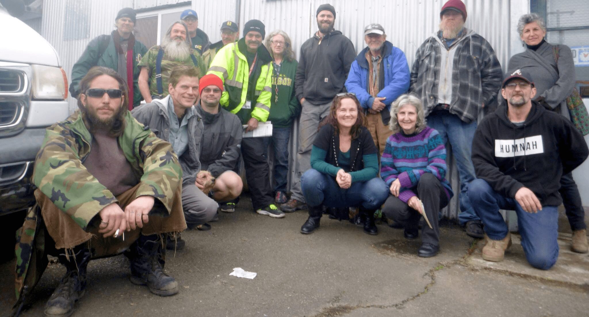 Group of people outdoors, some sitting and some standing, in front of a vehicle, smiling and posing for the photo.