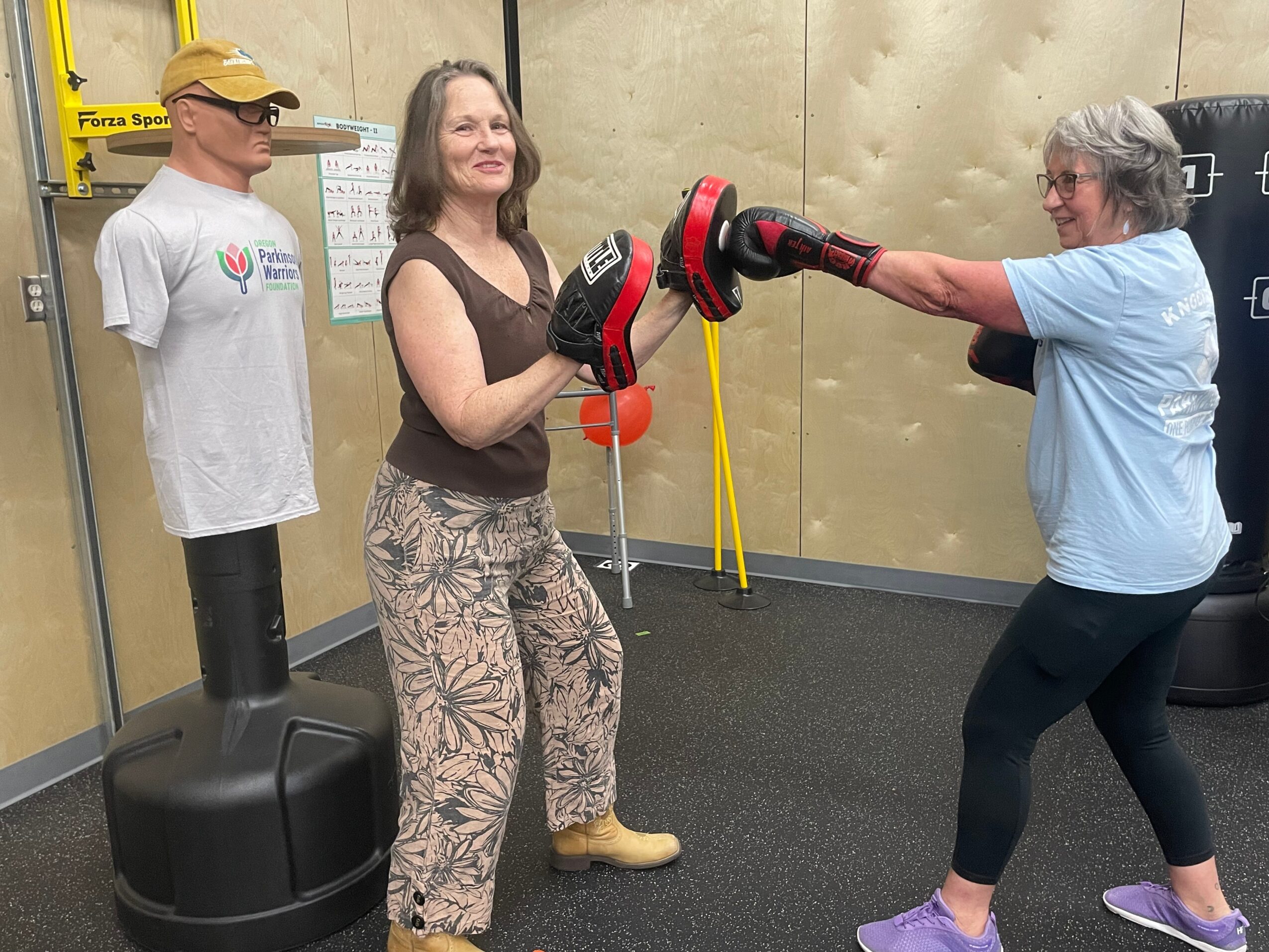 Two women practicing boxing with a punching mitt and a punching bag in a gym, a mannequin stands nearby, and a yellow caution sign is visible.