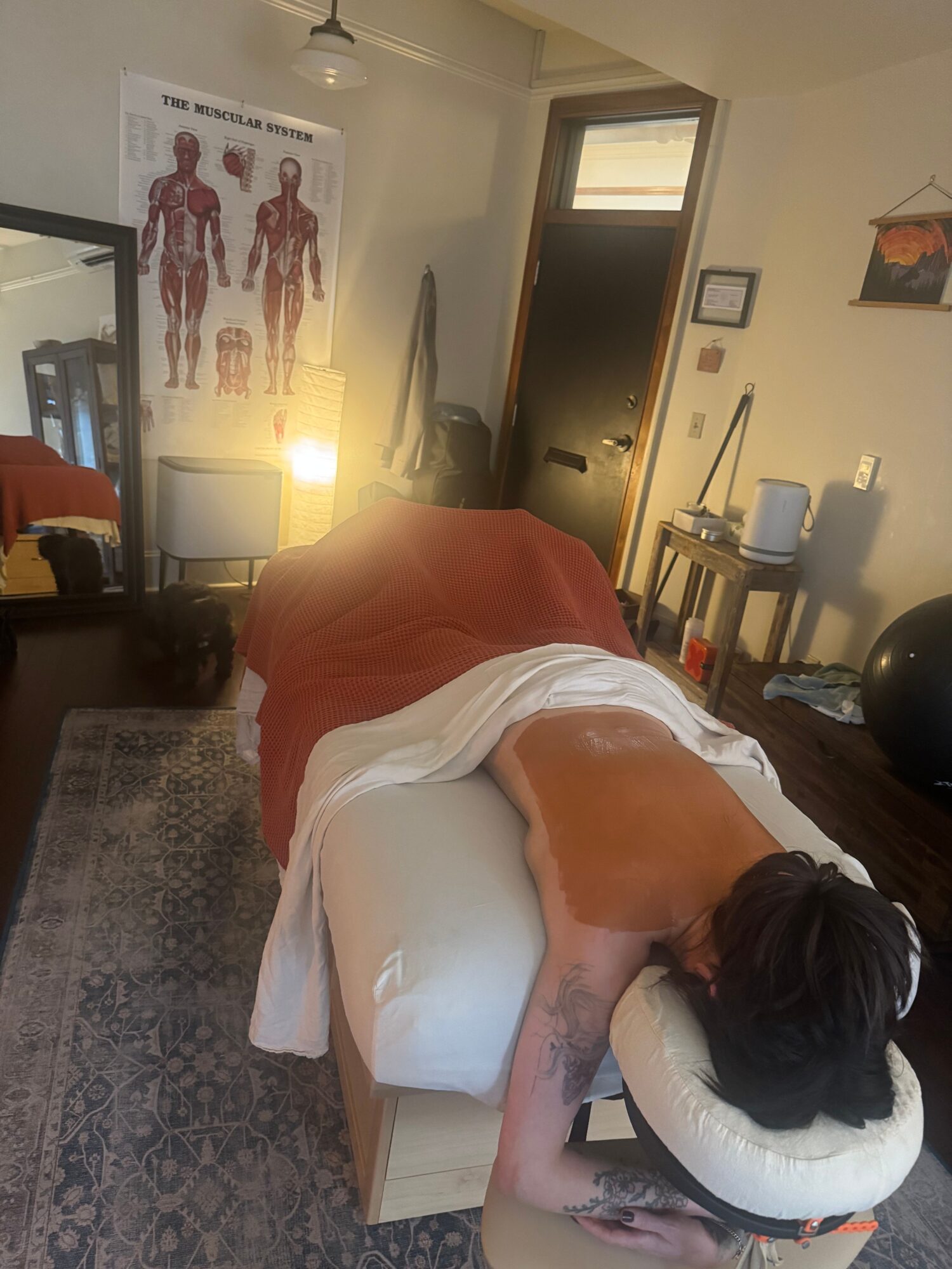 Person lying face down on a massage table with a towel, in a room with a door, mirror, and medical poster.