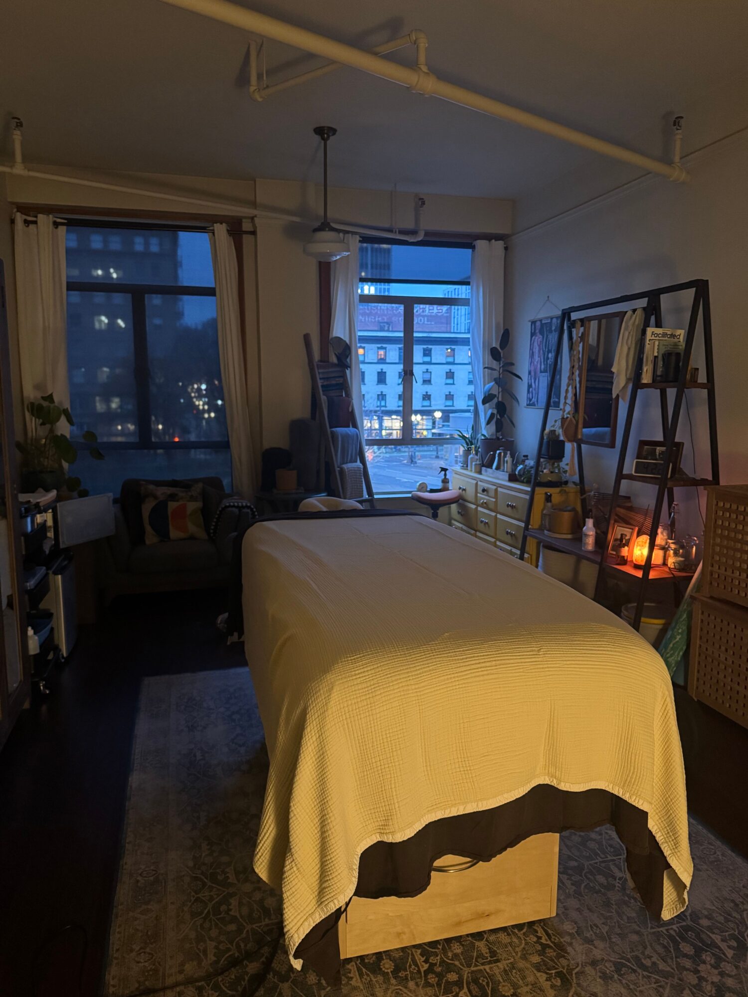 Bedroom with bed, window, bookshelf, and a small table with a candle, illuminated by evening light.