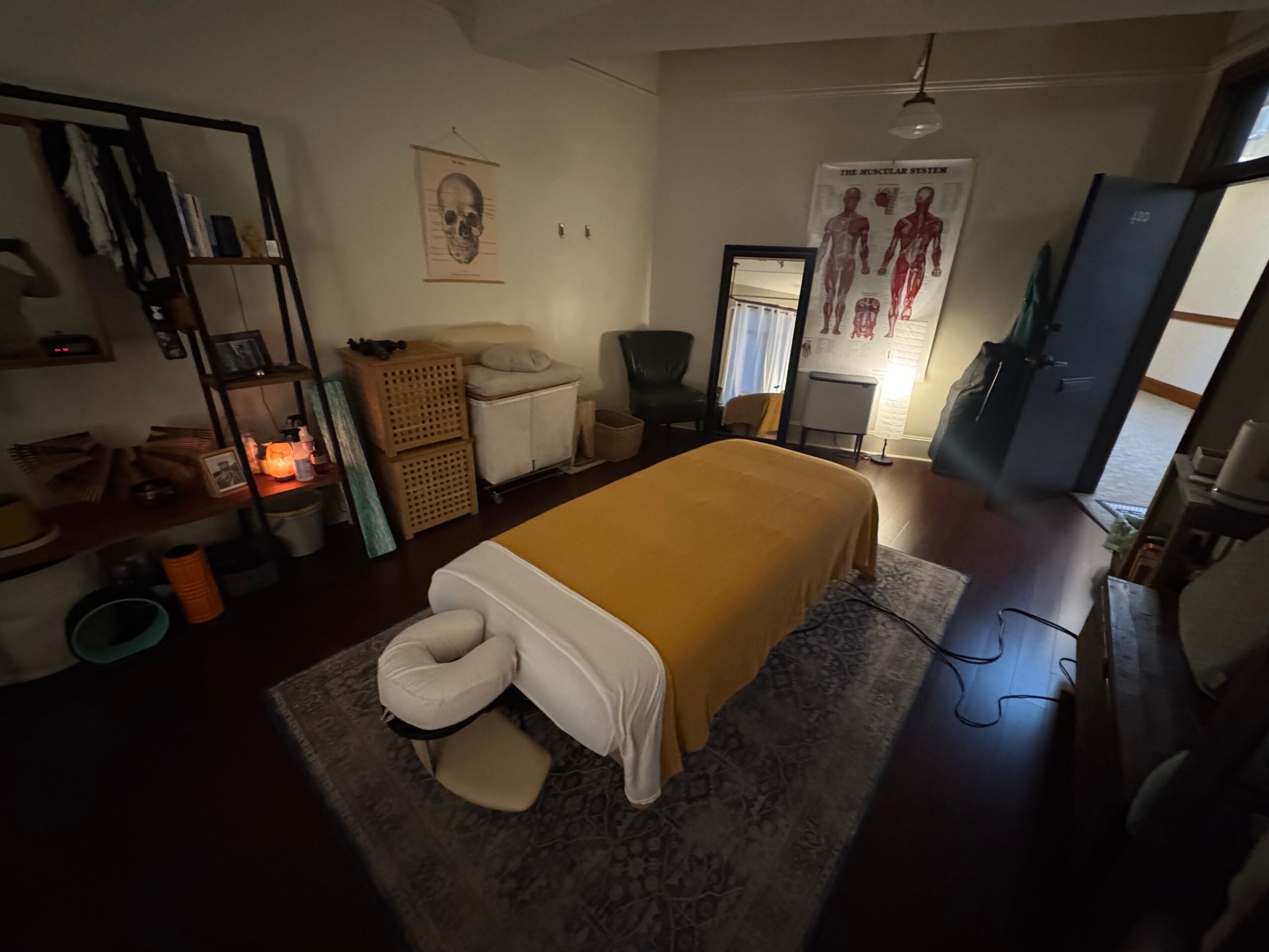 Room with massage table covered by yellow sheet, surrounded by furniture and artwork, with a dark wooden floor and window.