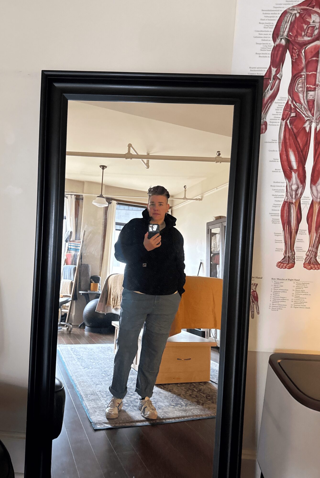 Person taking a mirror selfie in a room with furniture and a large poster of human anatomy on the wall.