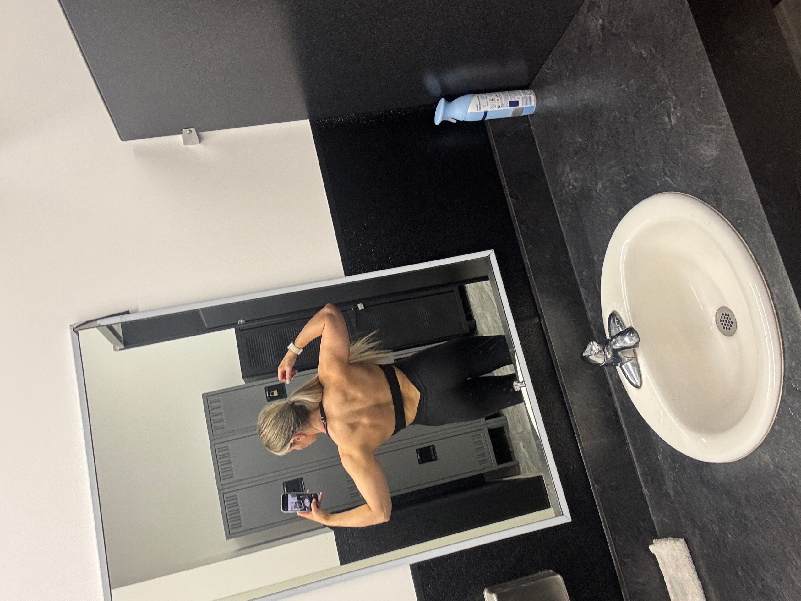 Person taking a mirror selfie in a locker room, shirtless, with lockers in the background, and a sink visible.