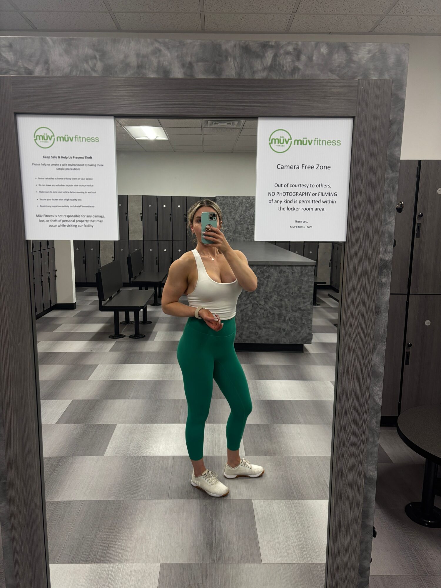 Person taking a mirror selfie in a gym, wearing white top and green pants, with a phone covering face.