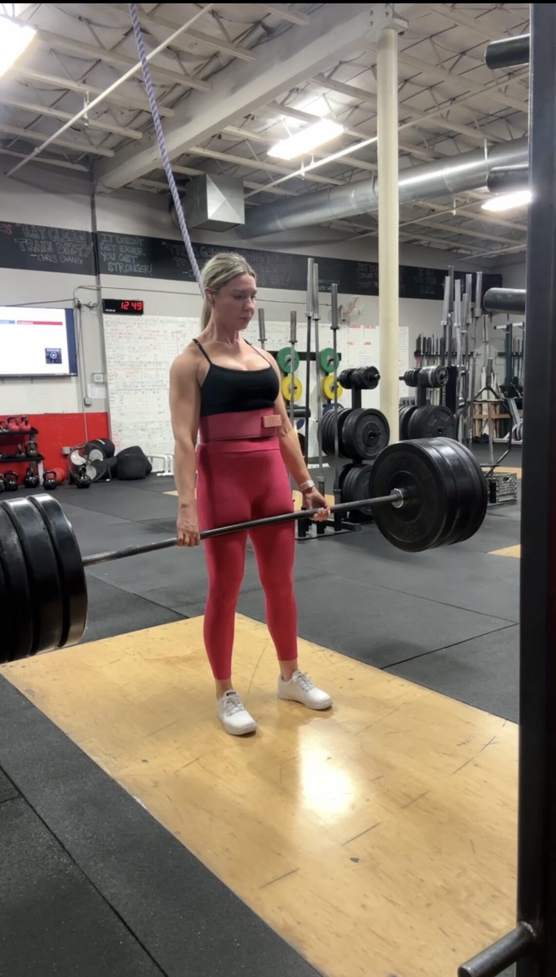 Woman in gym lifting a barbell with weights, standing on a wooden platform, gym equipment in background.