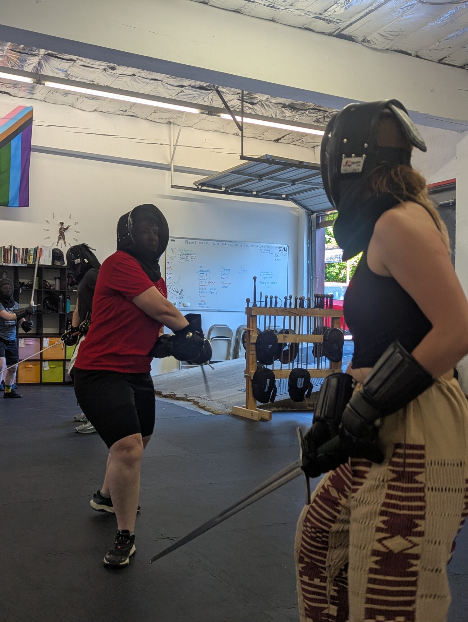 Two women in a gym, one in a red shirt and black shorts, holding a battle rope, the other in a helmet and gloves, standing nearby.