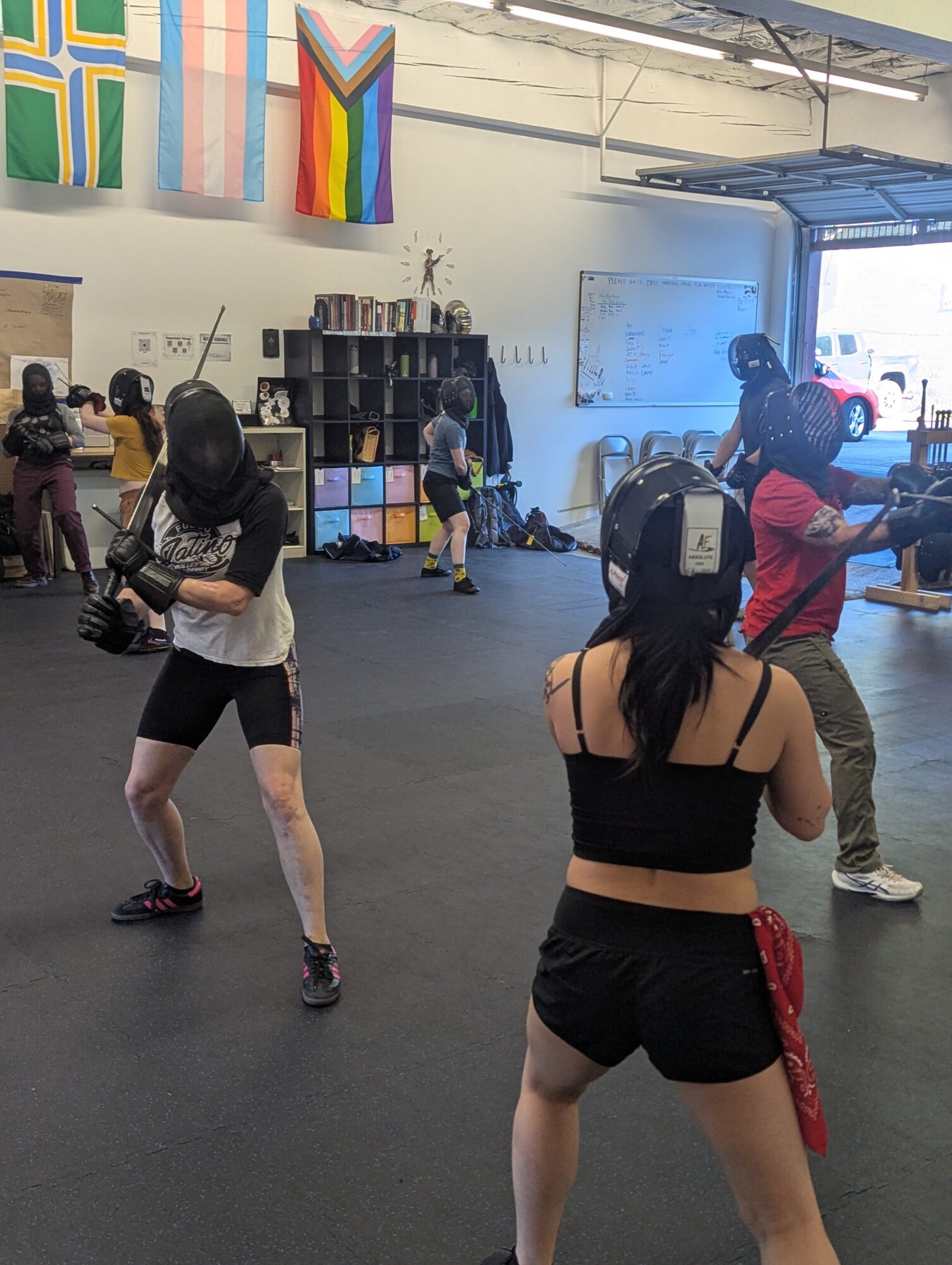 People practicing boxing in a gym with rainbow flags and a whiteboard, some wearing protective gear, others training.
