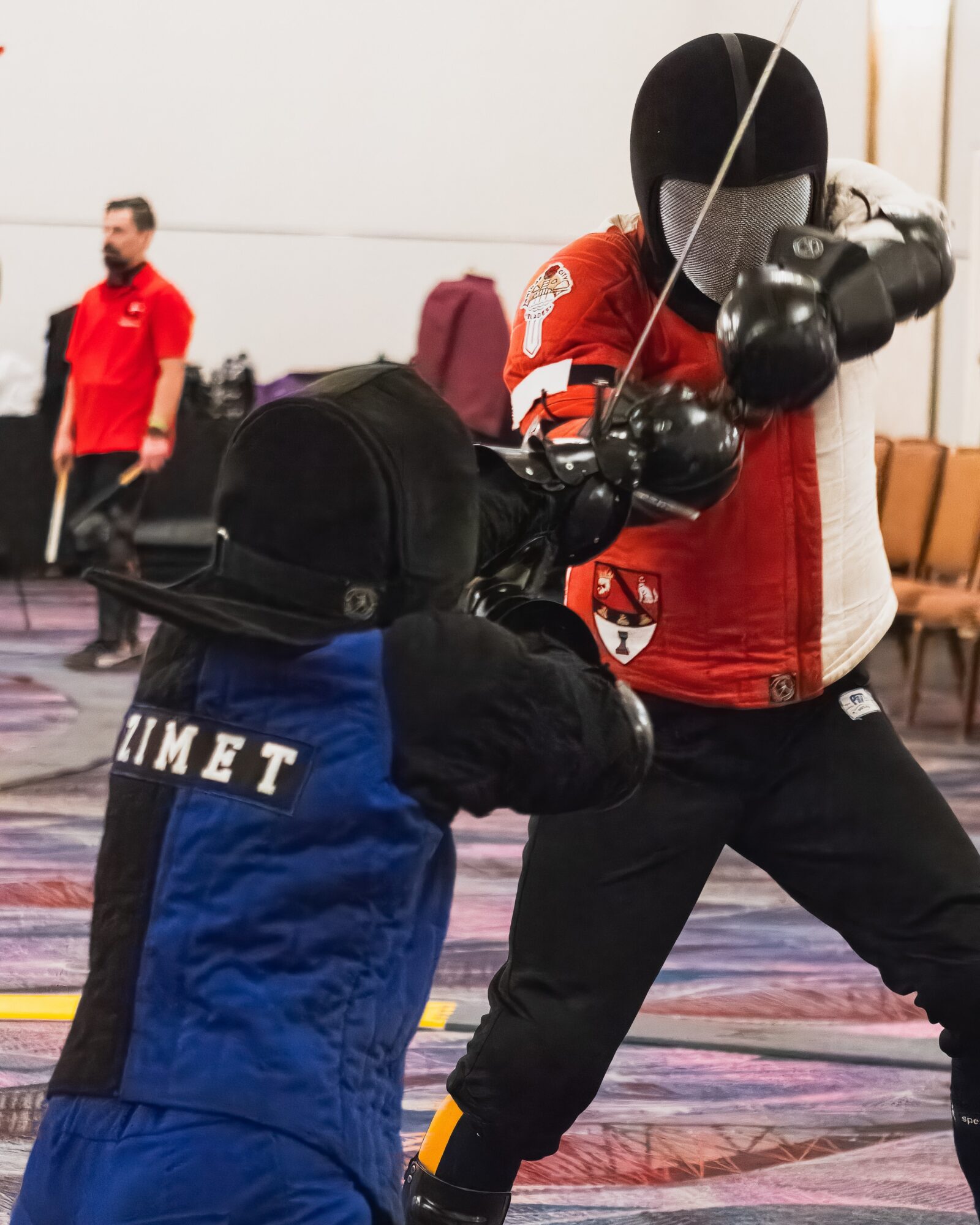 Person in red jacket practicing fencing with a mask and sword, in an indoor sports facility.