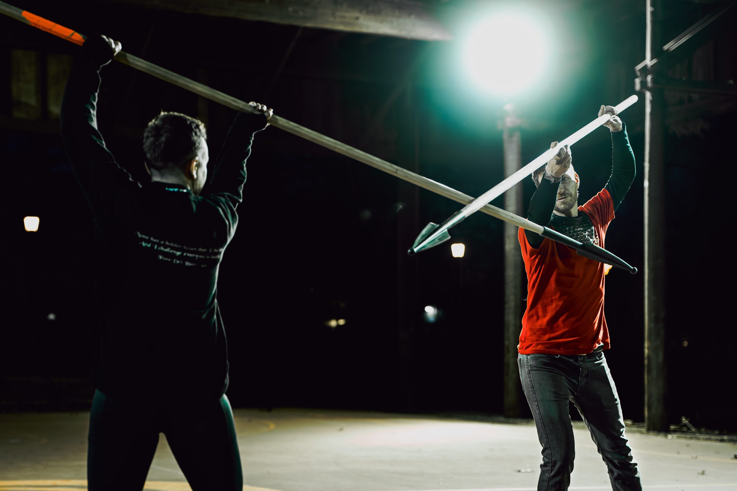 Two performers practicing with long poles in a dark indoor space, illuminated by bright overhead light.