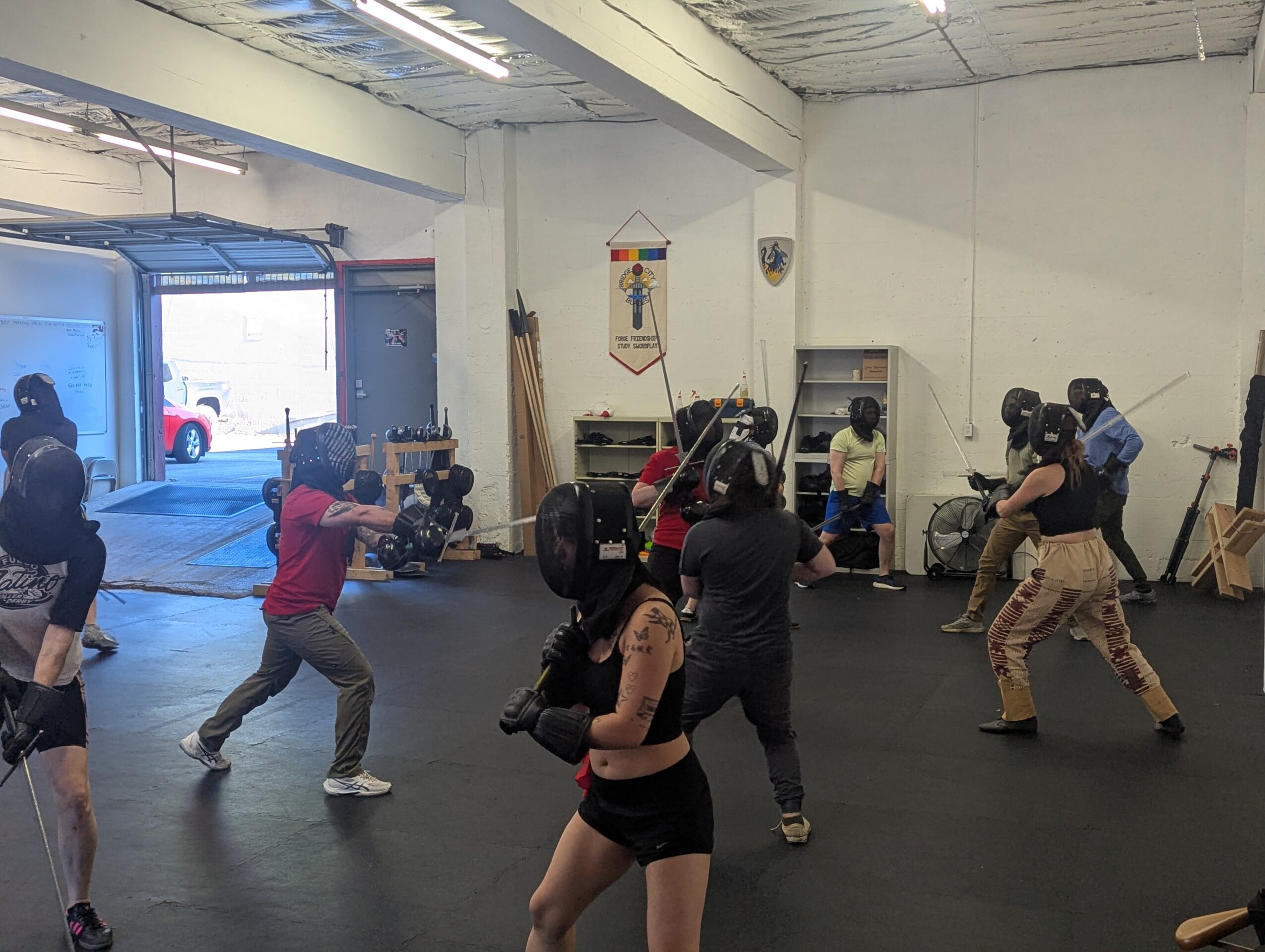 People practicing boxing in a gym, some wearing gloves and headgear, with a large open door showing outside view.