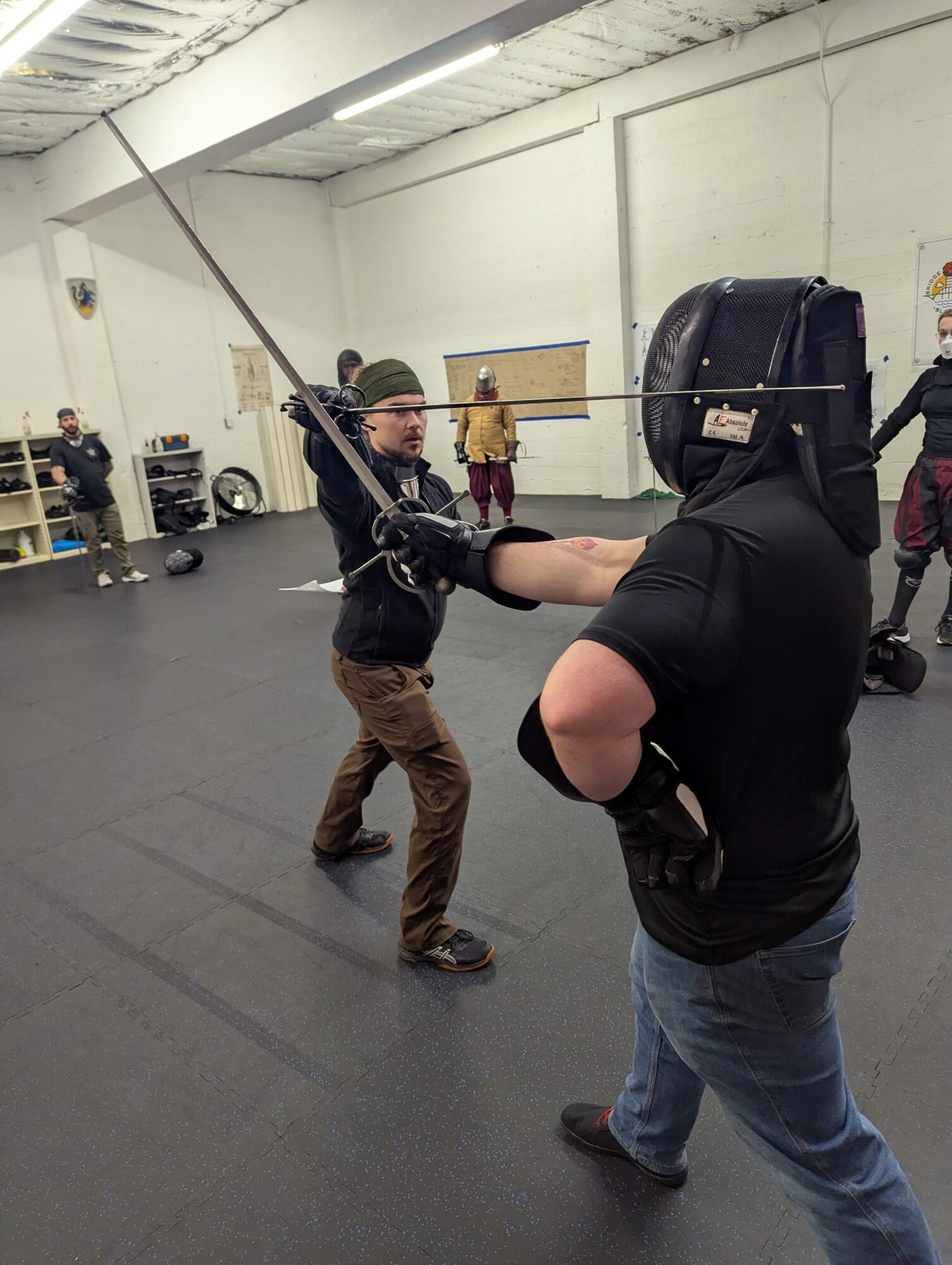 Person practicing sword fighting with another person in a gym, others observing in the background.