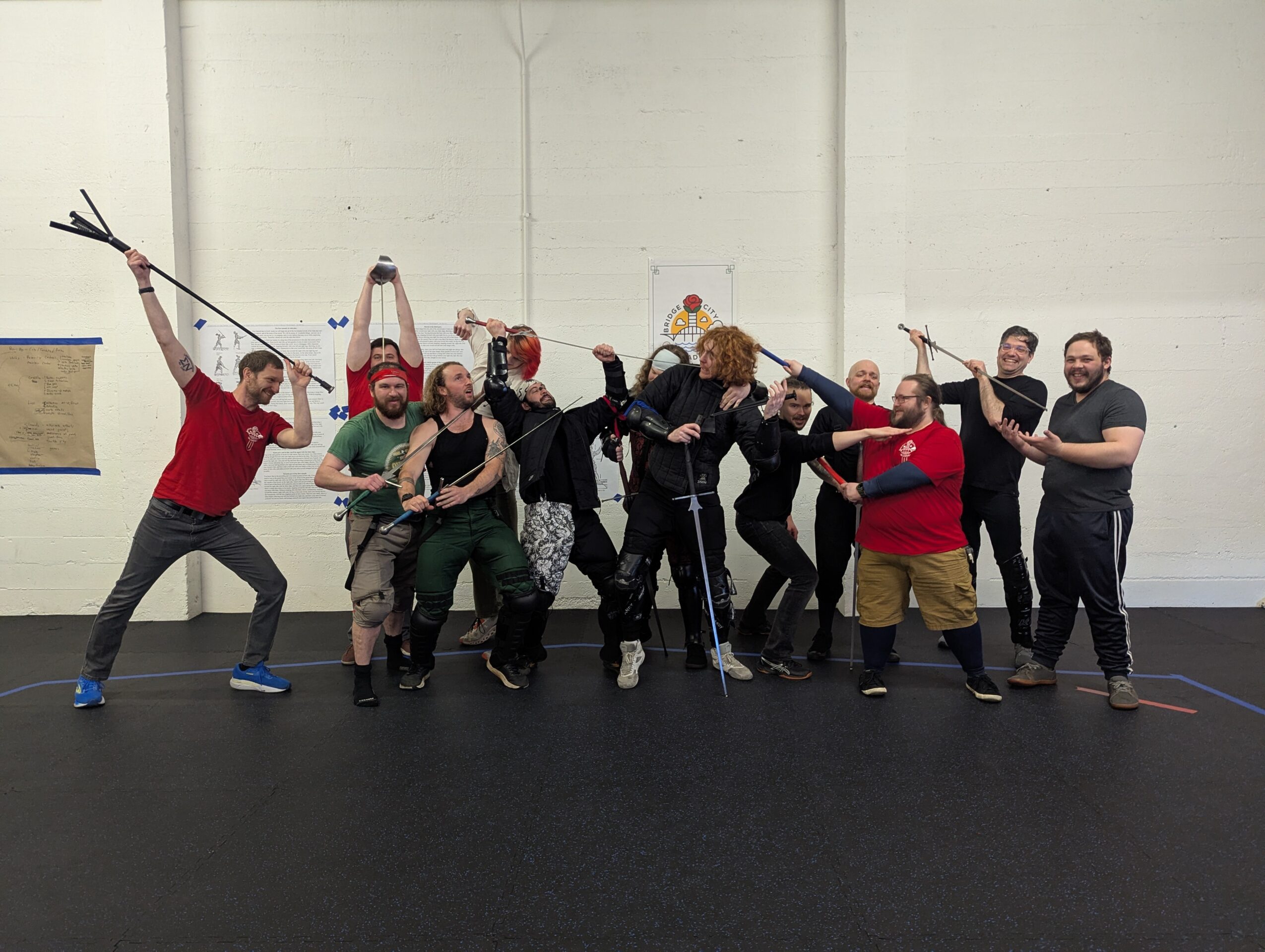 Group of people in a gym, holding sticks and posing playfully, some with arms raised, in front of a white wall.