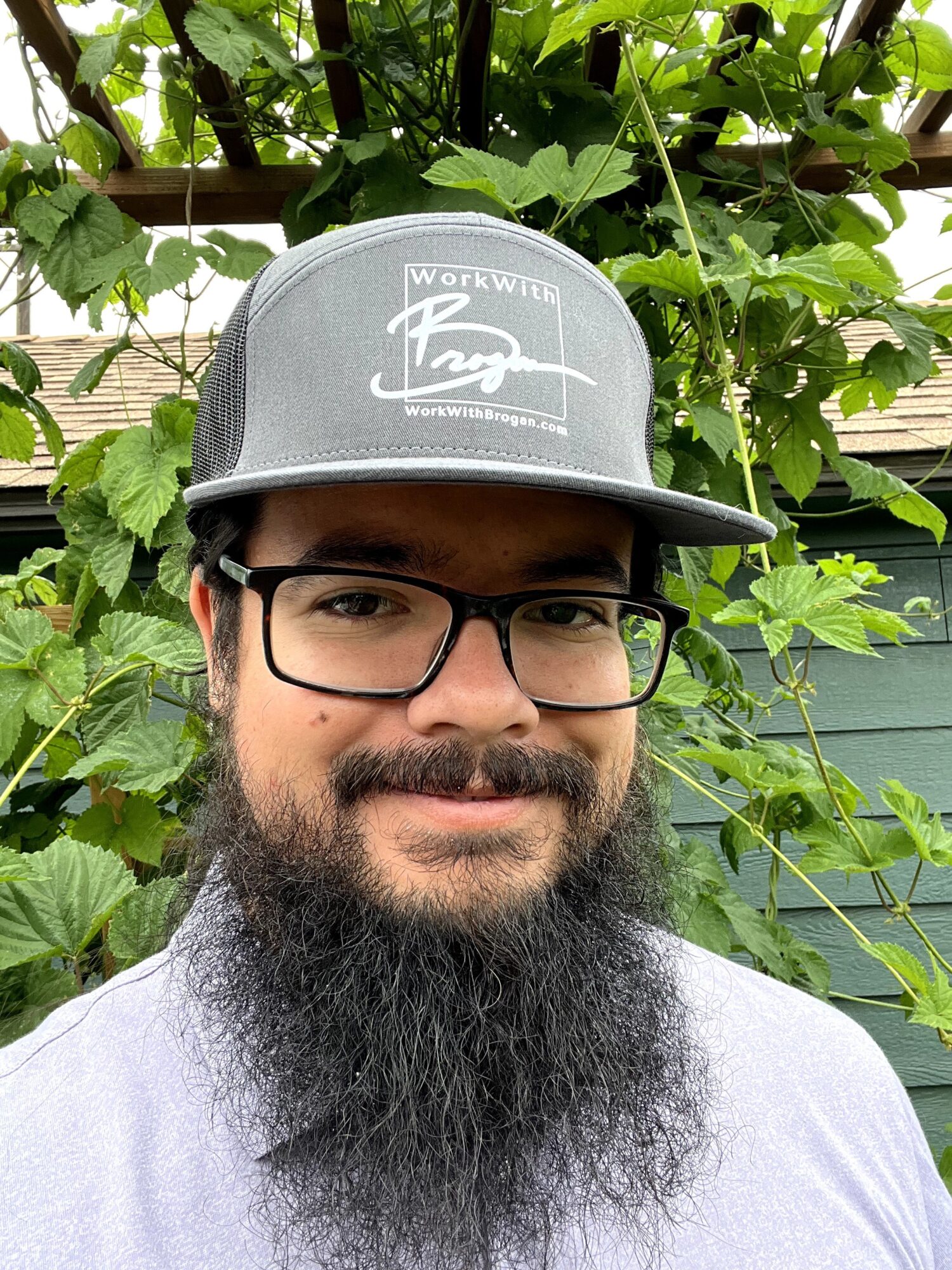 Man with glasses and a beard wearing a gray cap outdoors with green foliage in the background.