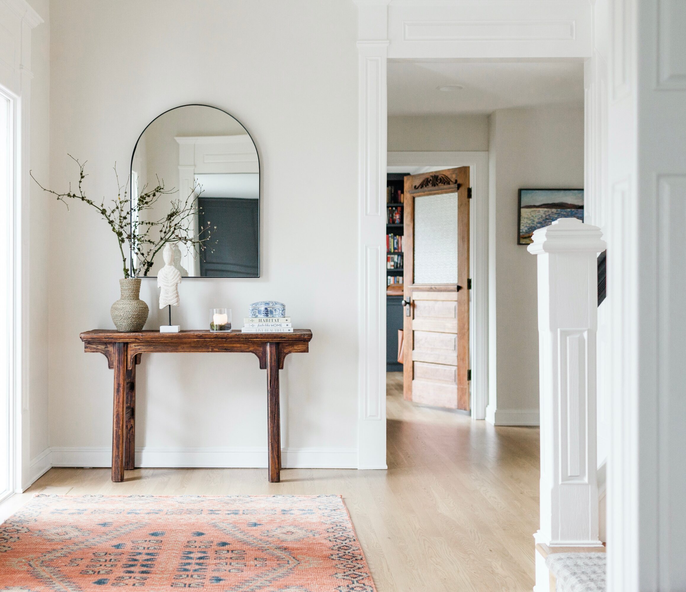 Entryway with a wooden console table, mirror, vase with branches, and a rug, leading to a room with a door and bookshelf.