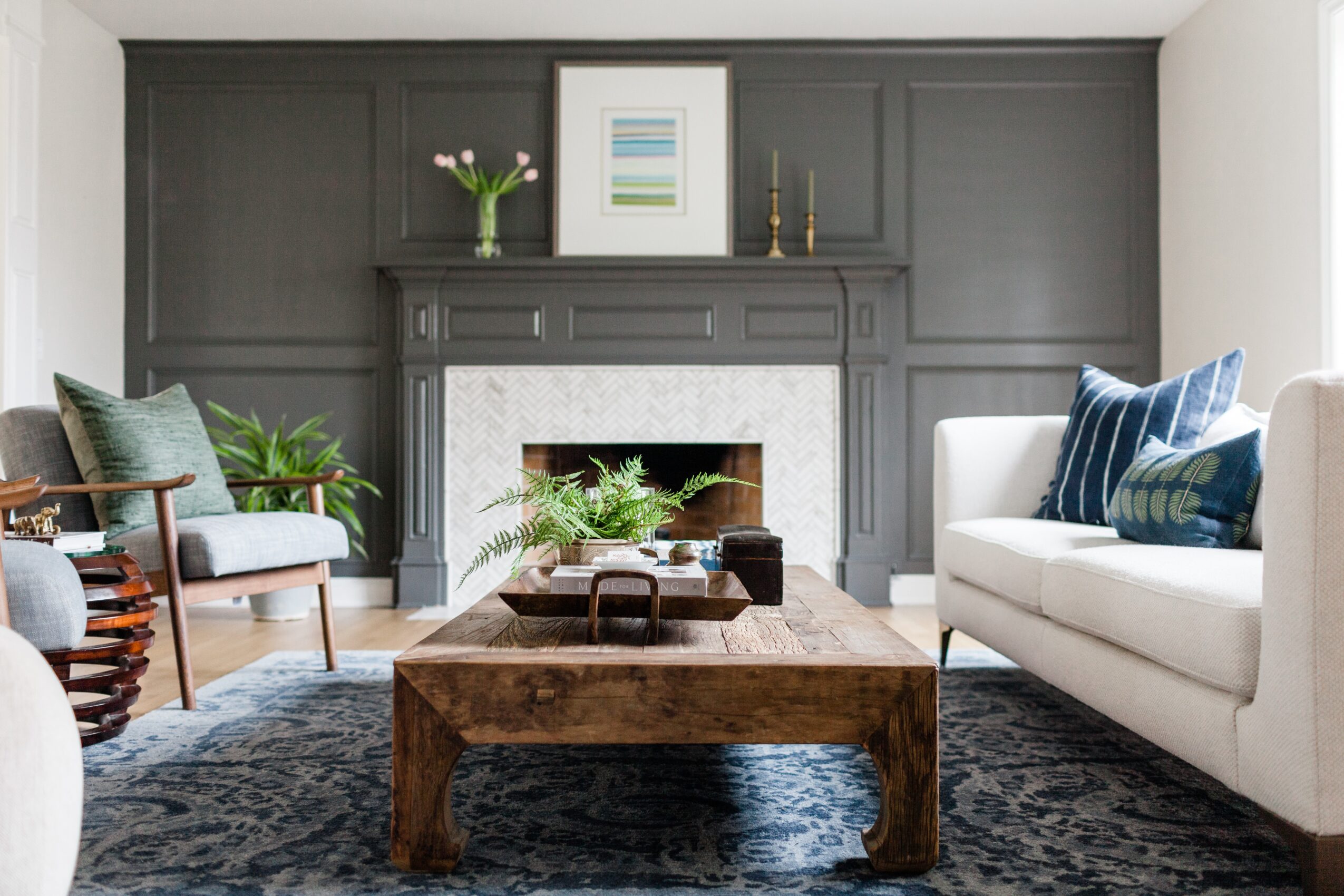 Living room with fireplace, gray wall paneling, white sofa with blue pillows, wooden coffee table, and plants.