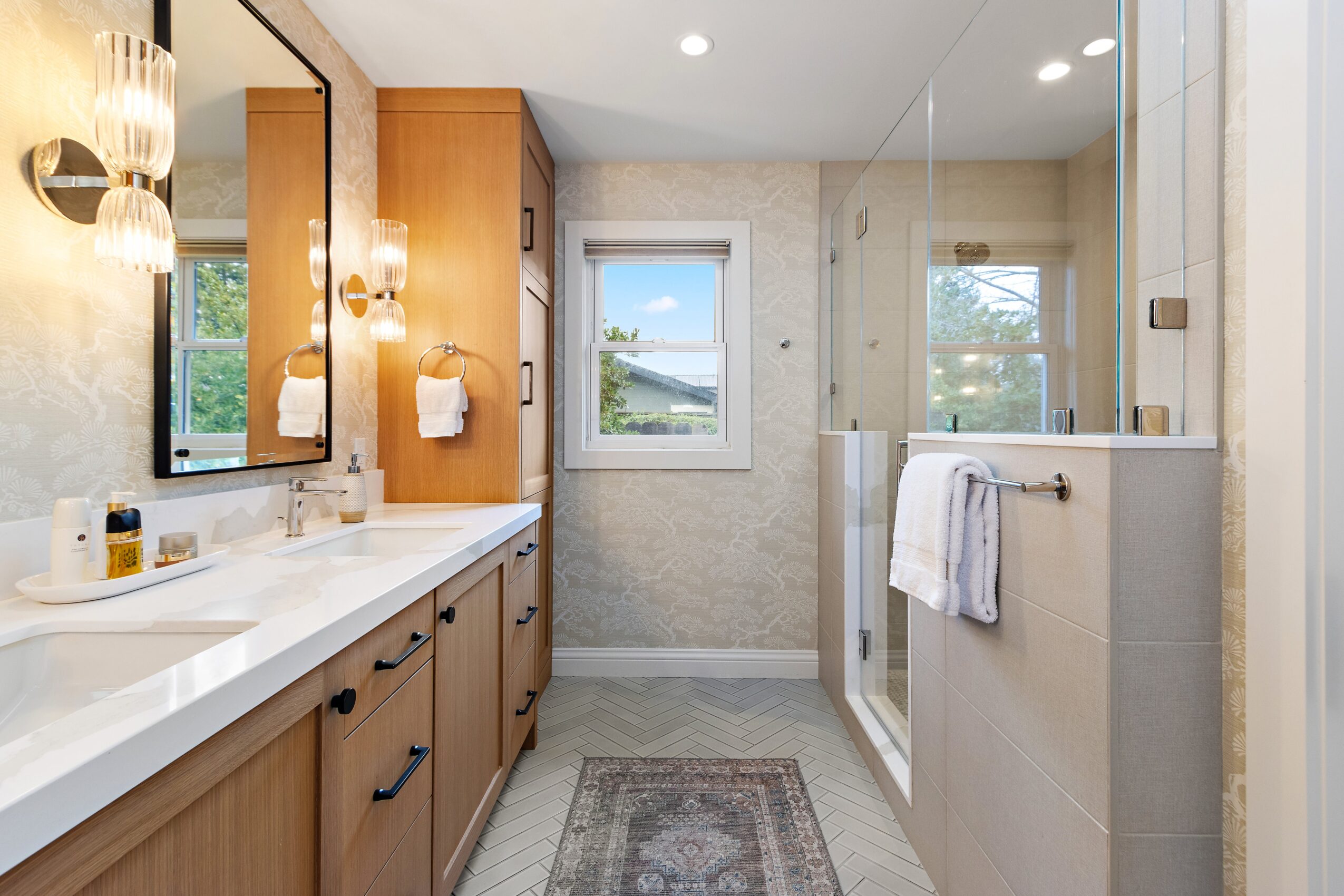 Bathroom with wooden cabinets, mirror, window, shower area, and towel rack, illuminated by wall lights and natural light.