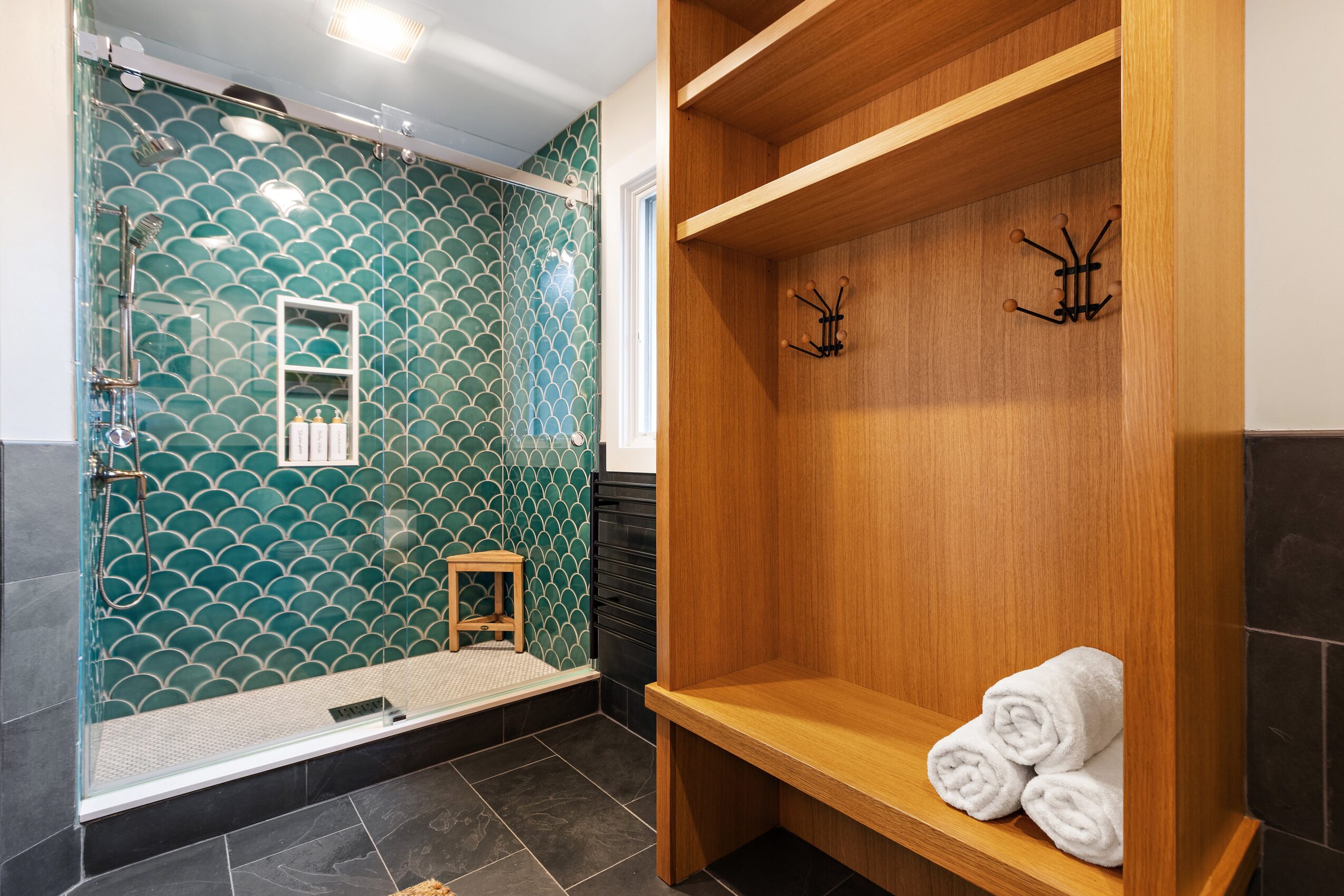 Bathroom with green tiled shower area, wooden shelf with rolled towels, and black and white flooring.