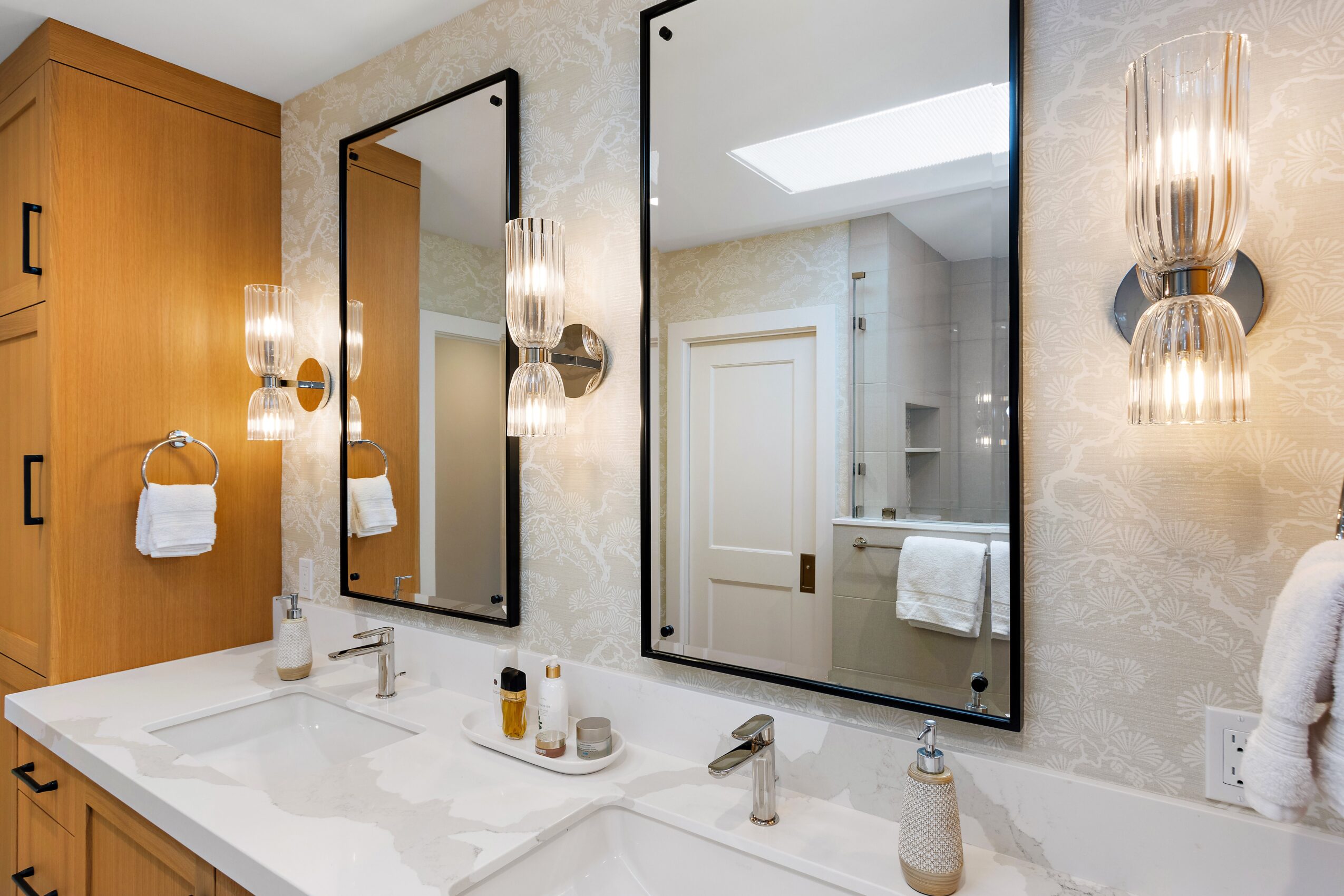 Bathroom with two large mirrors, wall-mounted lights, a white countertop, and wooden cabinets, reflected in the mirrors.
