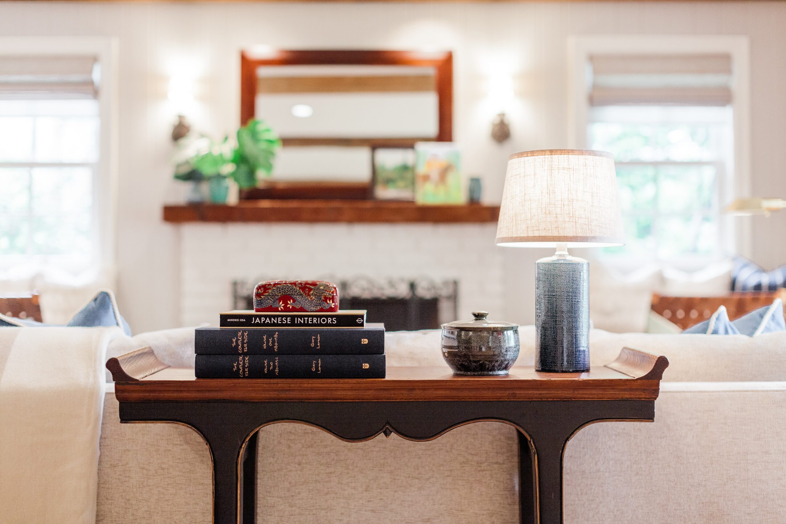 Living room with a wooden table, lamp, books, and decorative items, fireplace, and windows in the background.