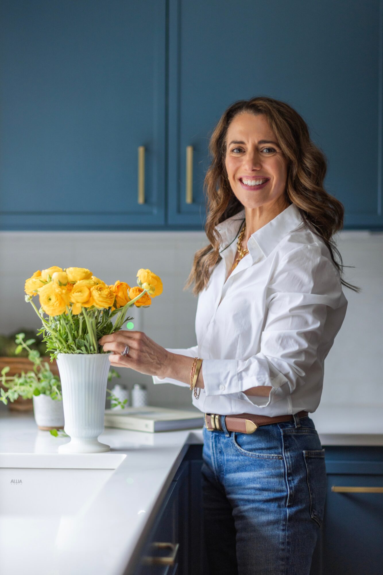 Woman with wavy hair smiling in a kitchen, holding a bouquet of yellow flowers in a white vase, wearing a white shirt and jeans.