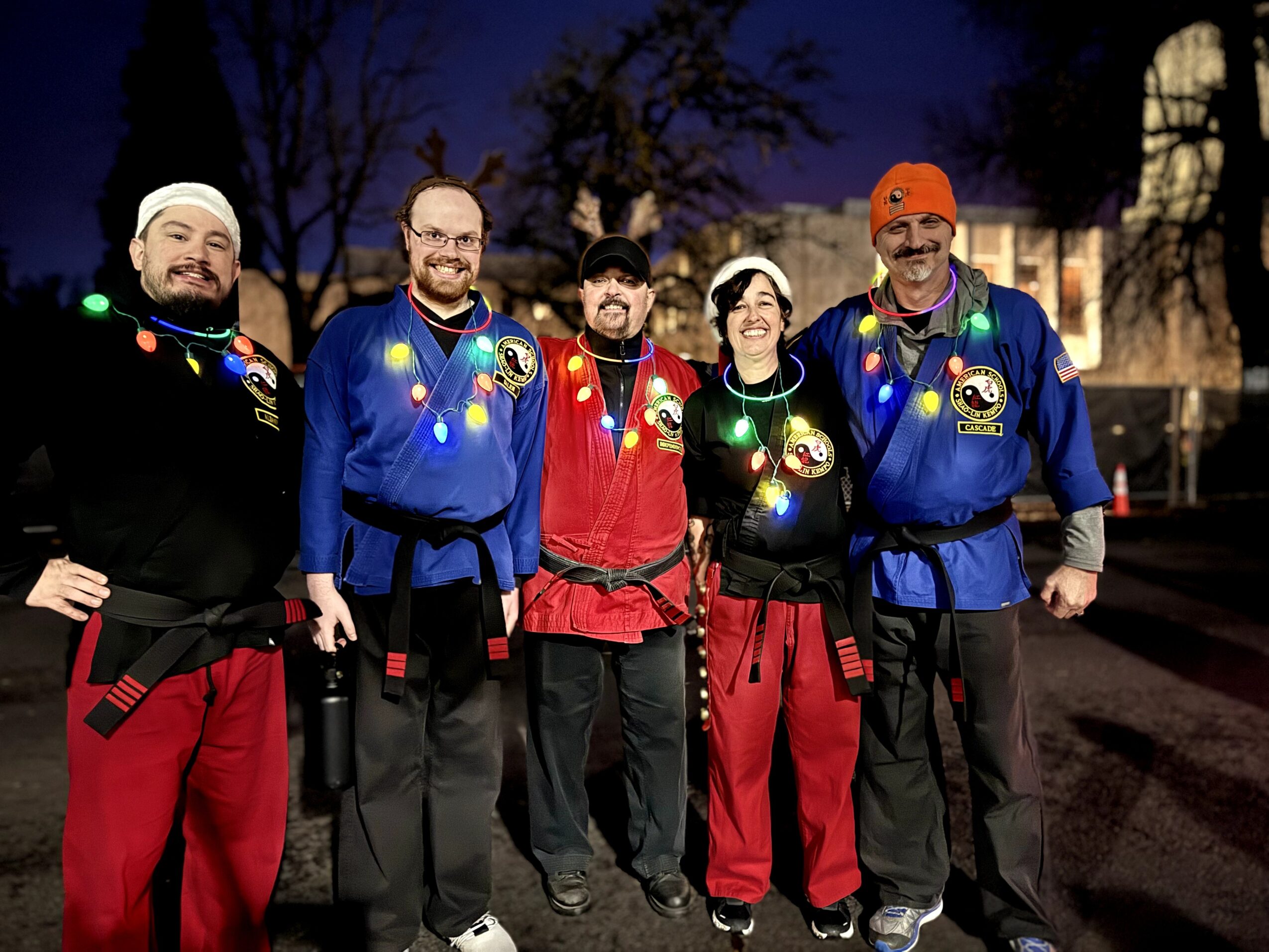 Five people dressed in colorful costumes with Christmas lights, standing outdoors at night, smiling.