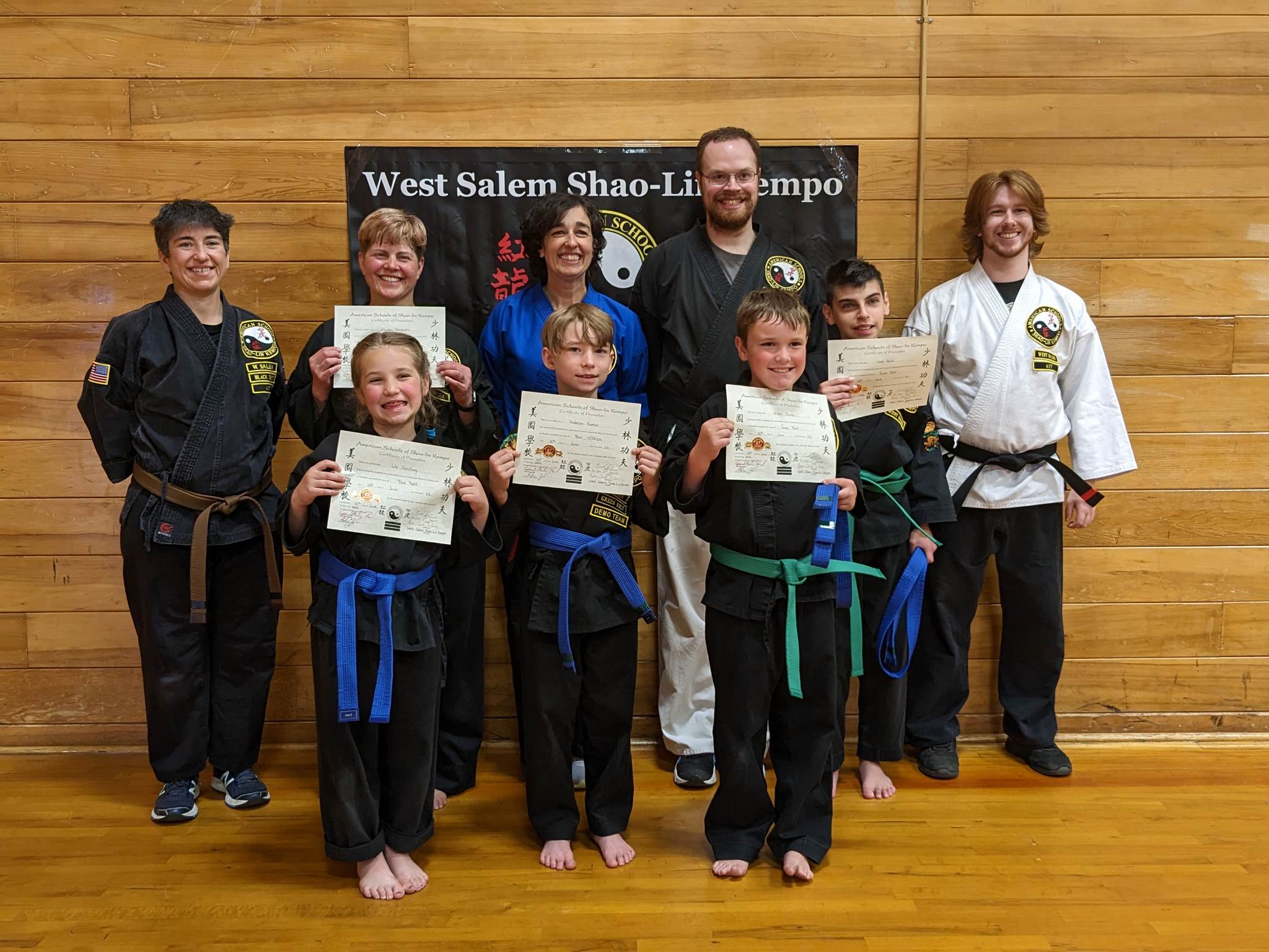 Group of children and instructors in martial arts uniforms holding certificates, standing on a wooden floor in front of a wooden wall and a sign.