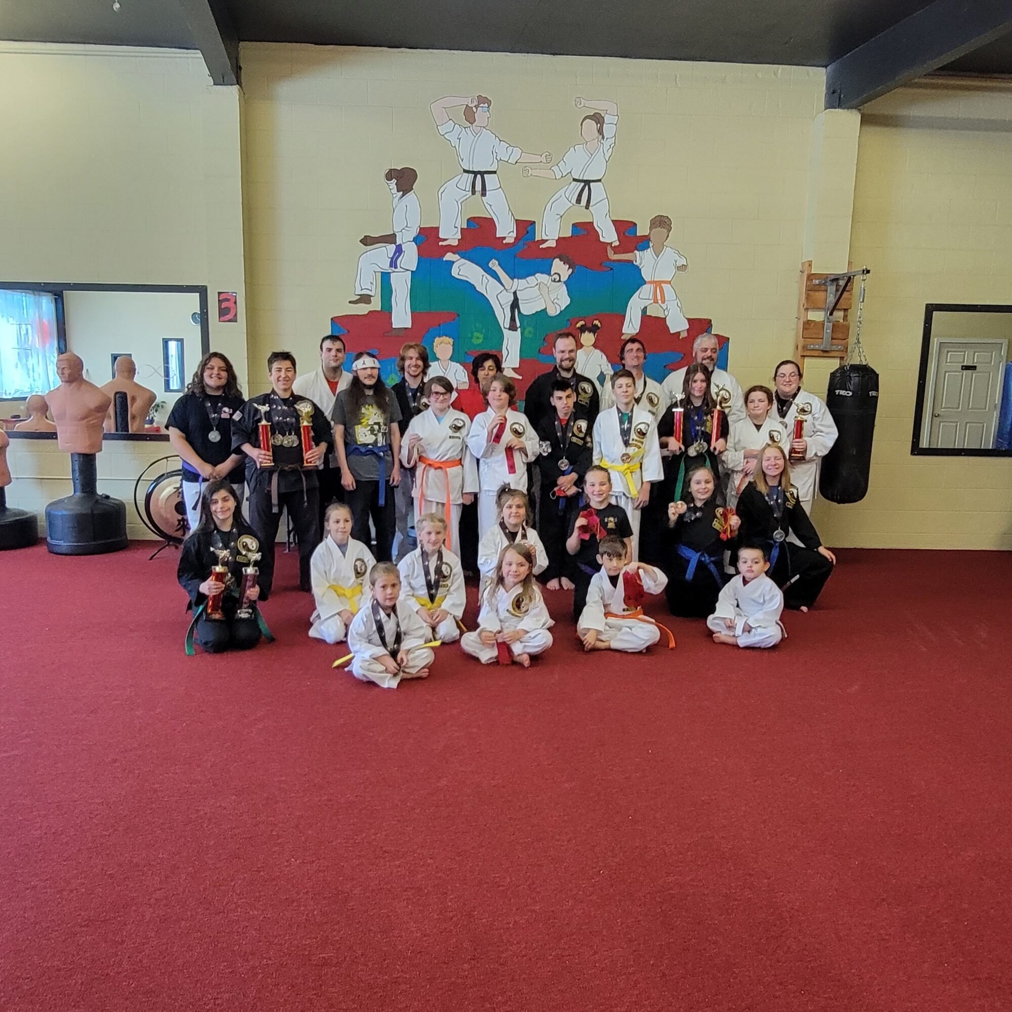 Group of children and adults sitting and standing on a red carpeted floor in a room with a yellow wall and a mural of cartoon characters. 20 words.