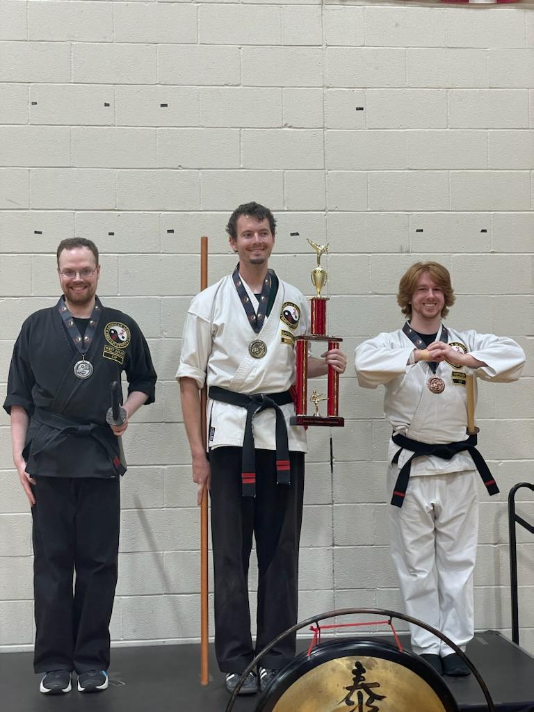 Three martial artists in uniforms with medals, standing indoors against a plain wall, one holding a large trophy.