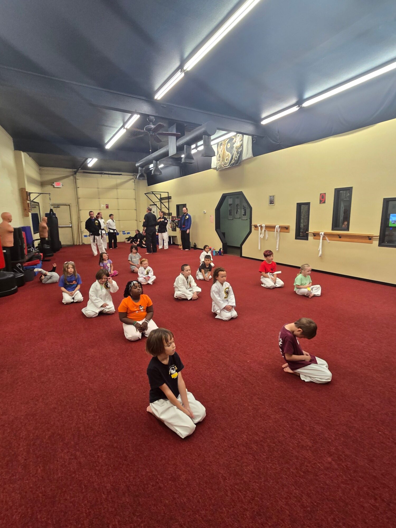 Children sitting on a red carpet in a room, some with adults, engaging in activities or conversations.