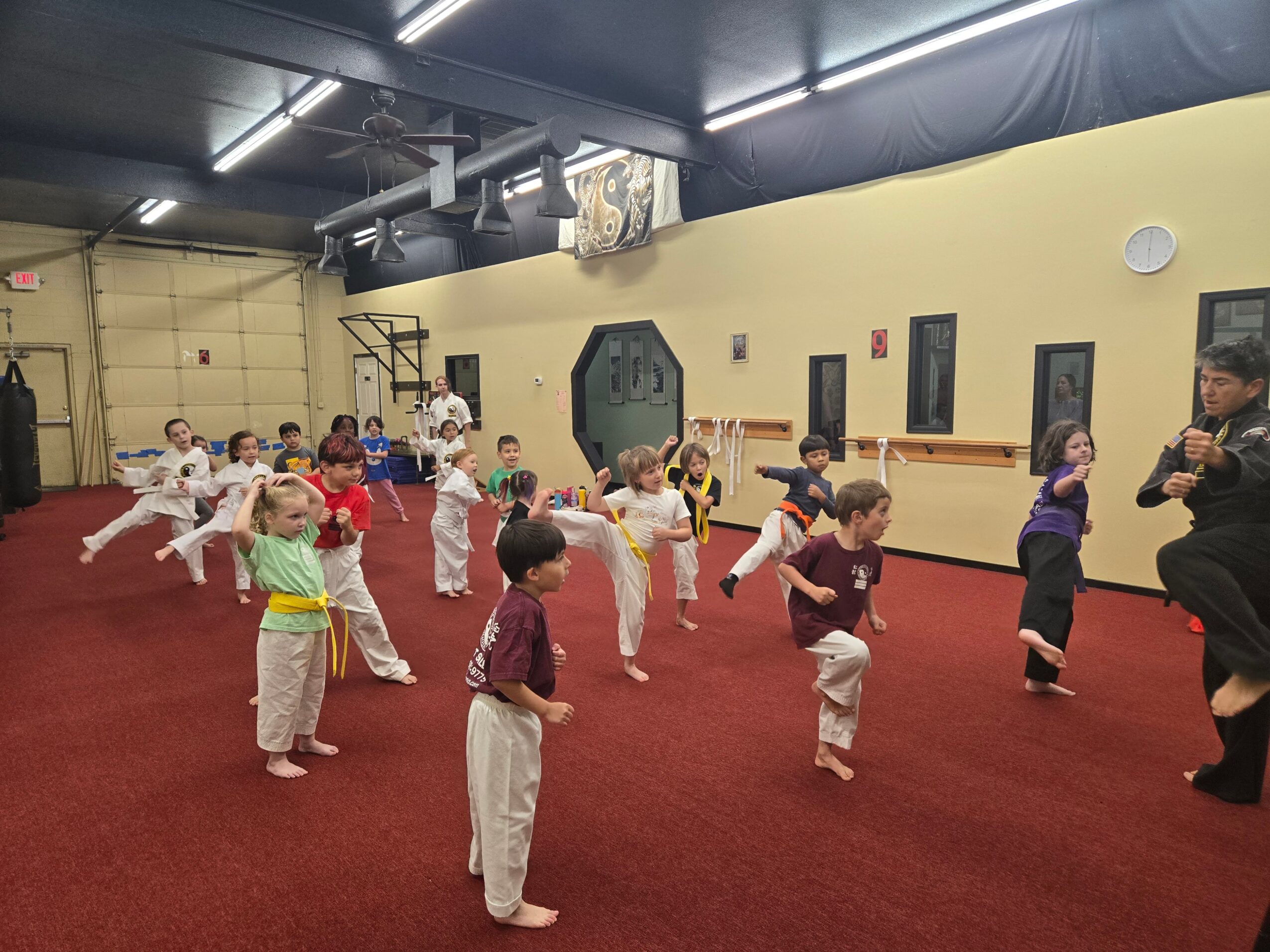 Children in martial arts uniforms practicing kicks in a gym with red carpeted floor and yellow walls.