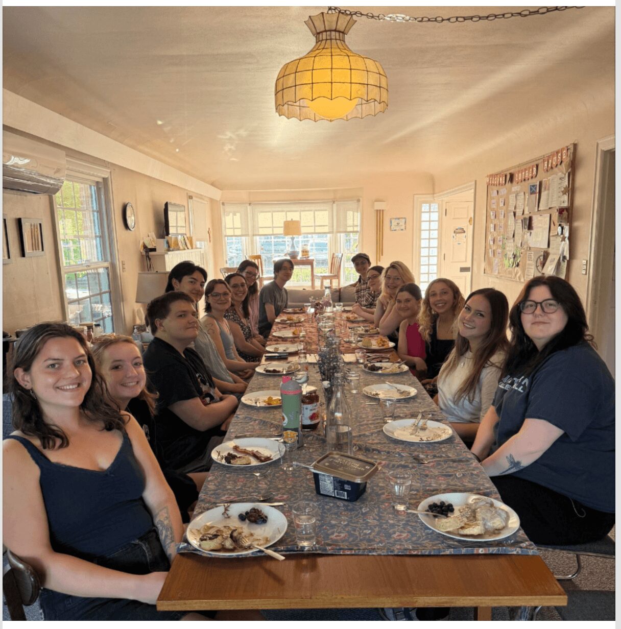 Group of people sitting at a long dining table in a bright room, smiling, with plates of food in front of them.