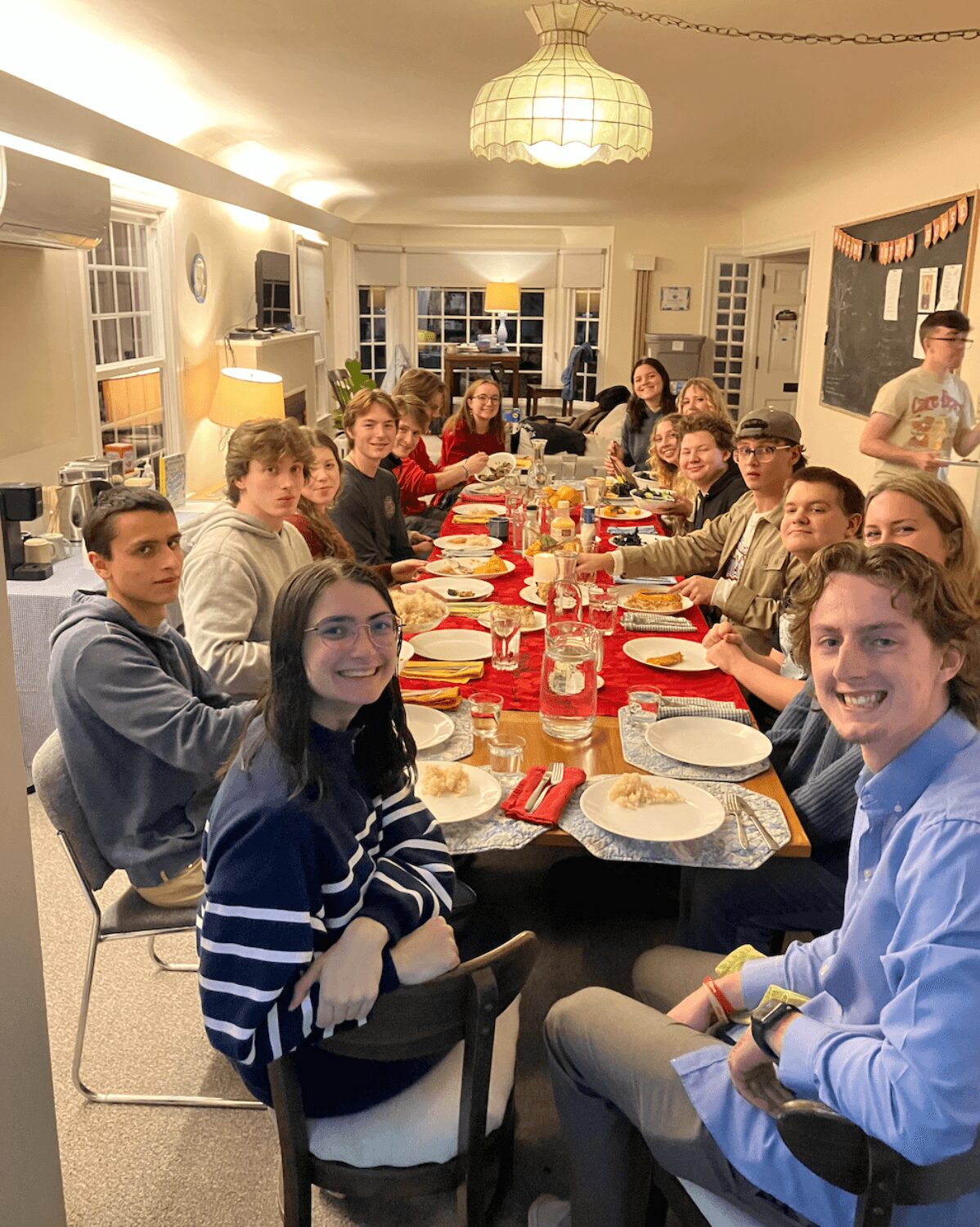 Group of people gathered around a long dining table with food and drinks in a well-lit room, smiling and enjoying a meal.
