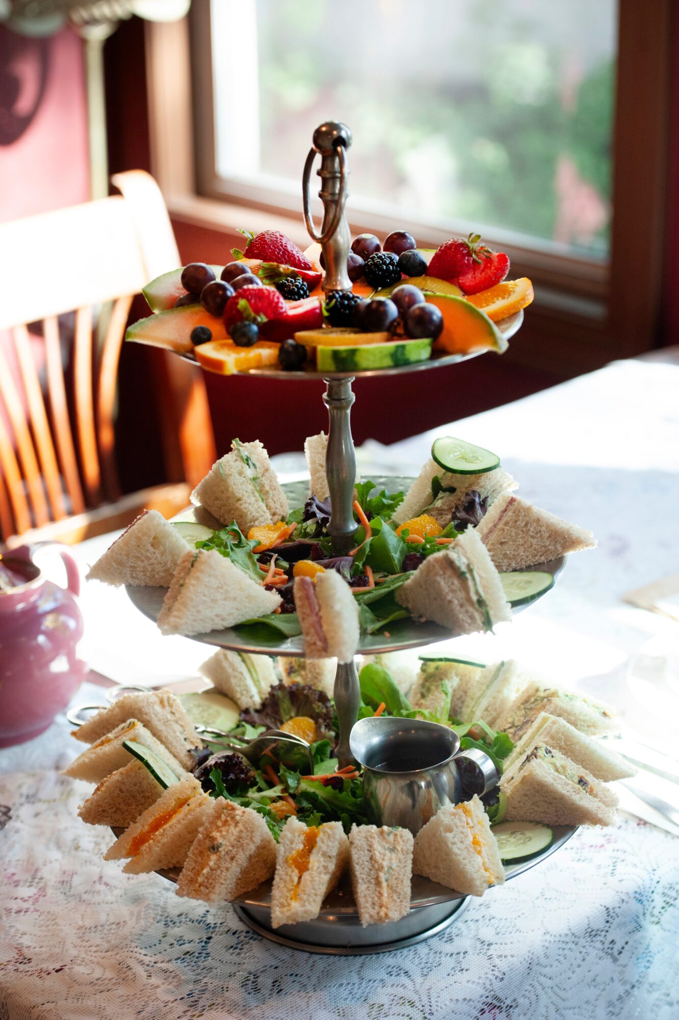 Three-tiered serving tray with sandwiches, fruits, and snacks on a table near a window, sunlight illuminating the scene.