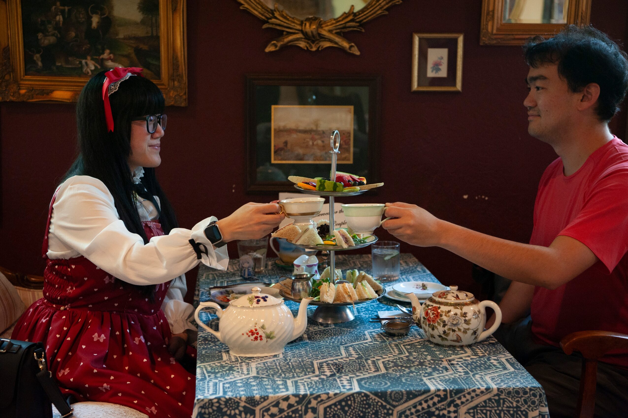 Two people sharing a tiered cake stand at a table with teapots and cups, indoors with framed pictures on the wall.
