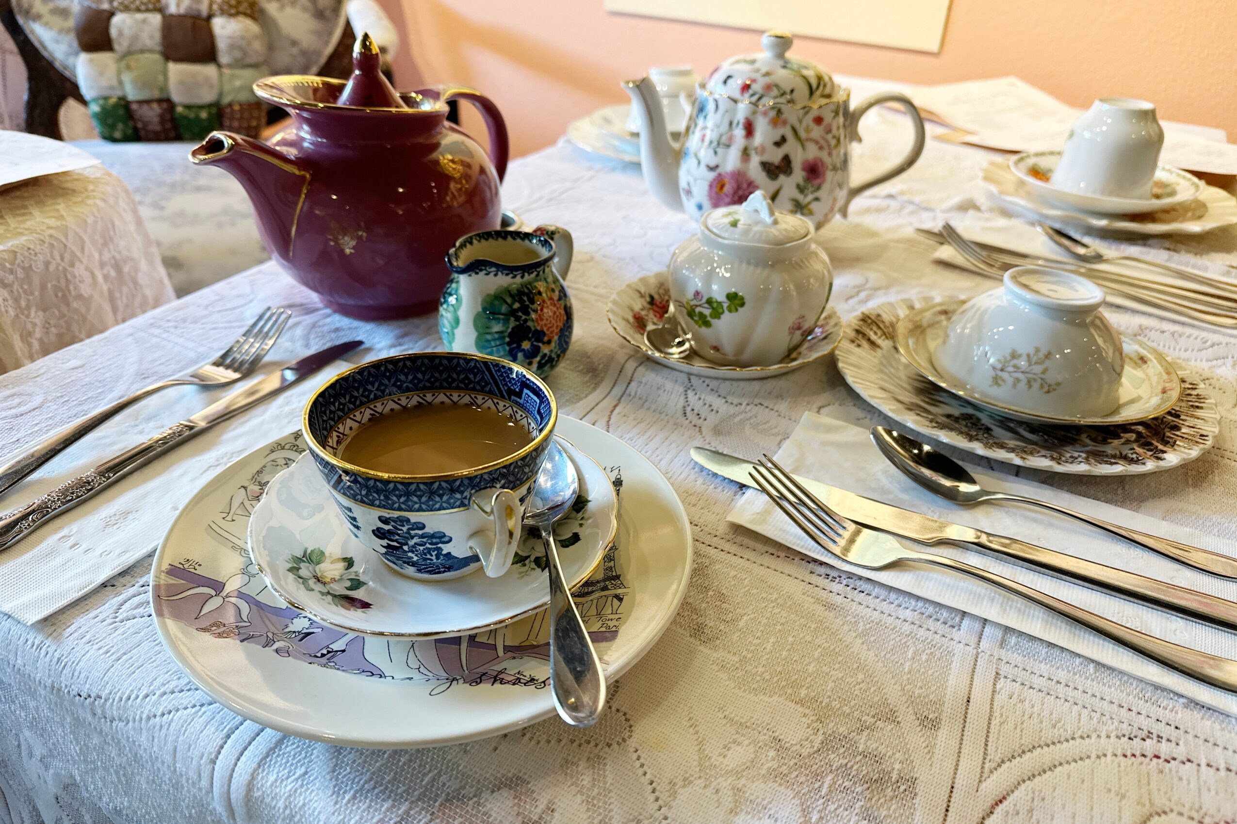 Tea set with cups, teapots, and plates on a tablecloth, with a cup of tea in the foreground, silverware, and decorative items.
