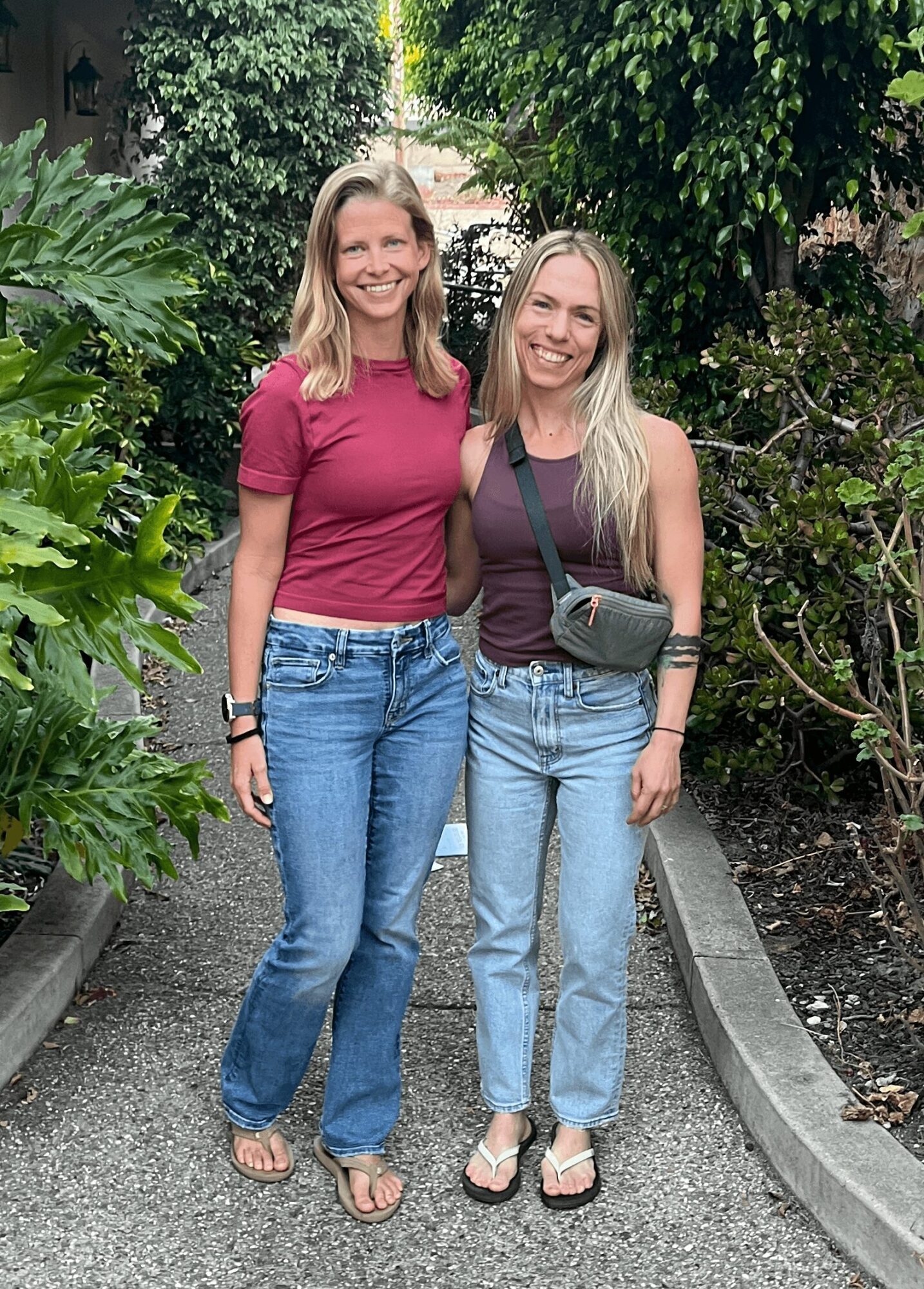 Two women stand outdoors on a garden path, smiling, surrounded by green plants and trees.