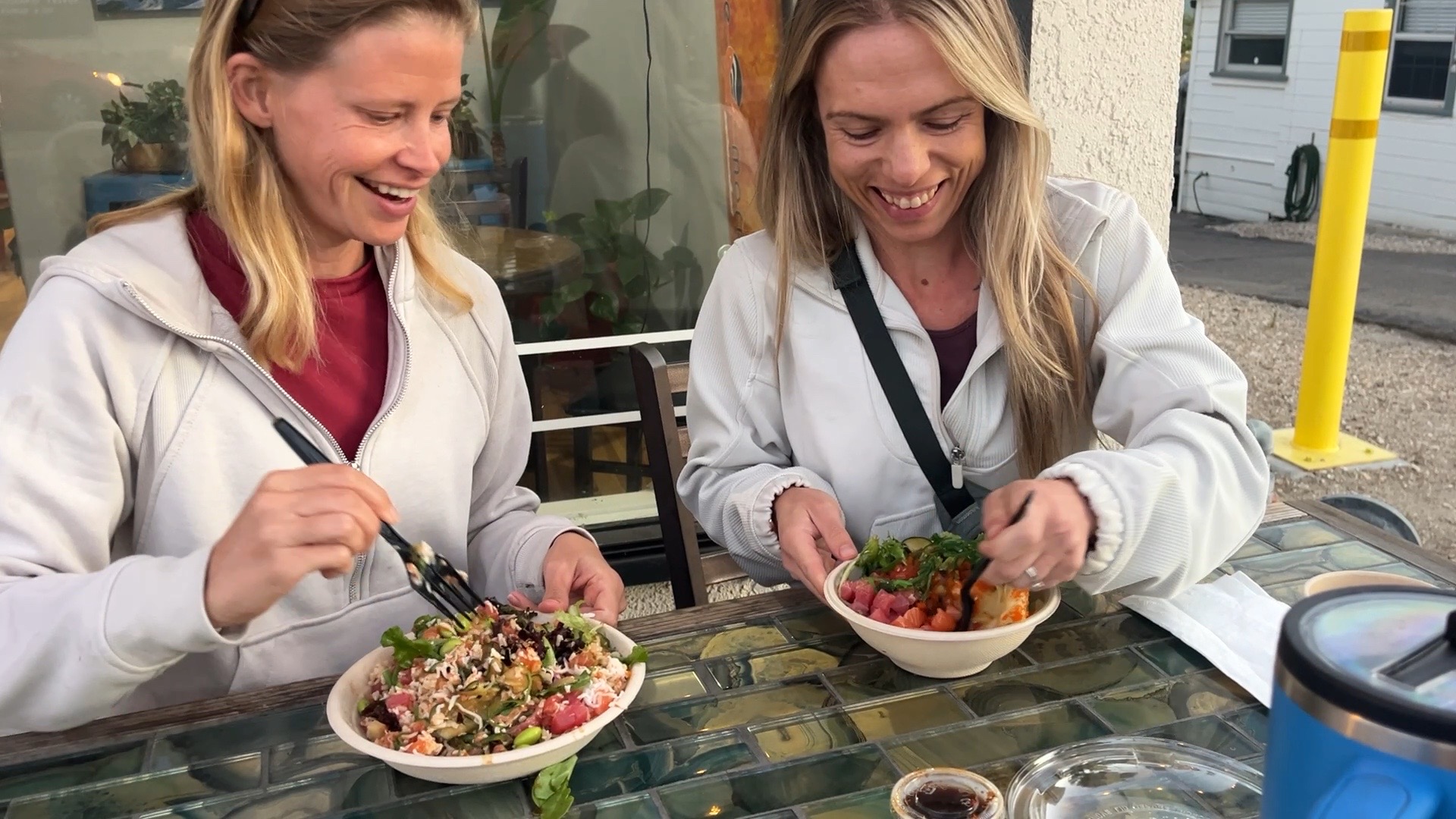 Two women sit at a table outdoors, smiling and eating salads from bowls, with a yellow post in the background.