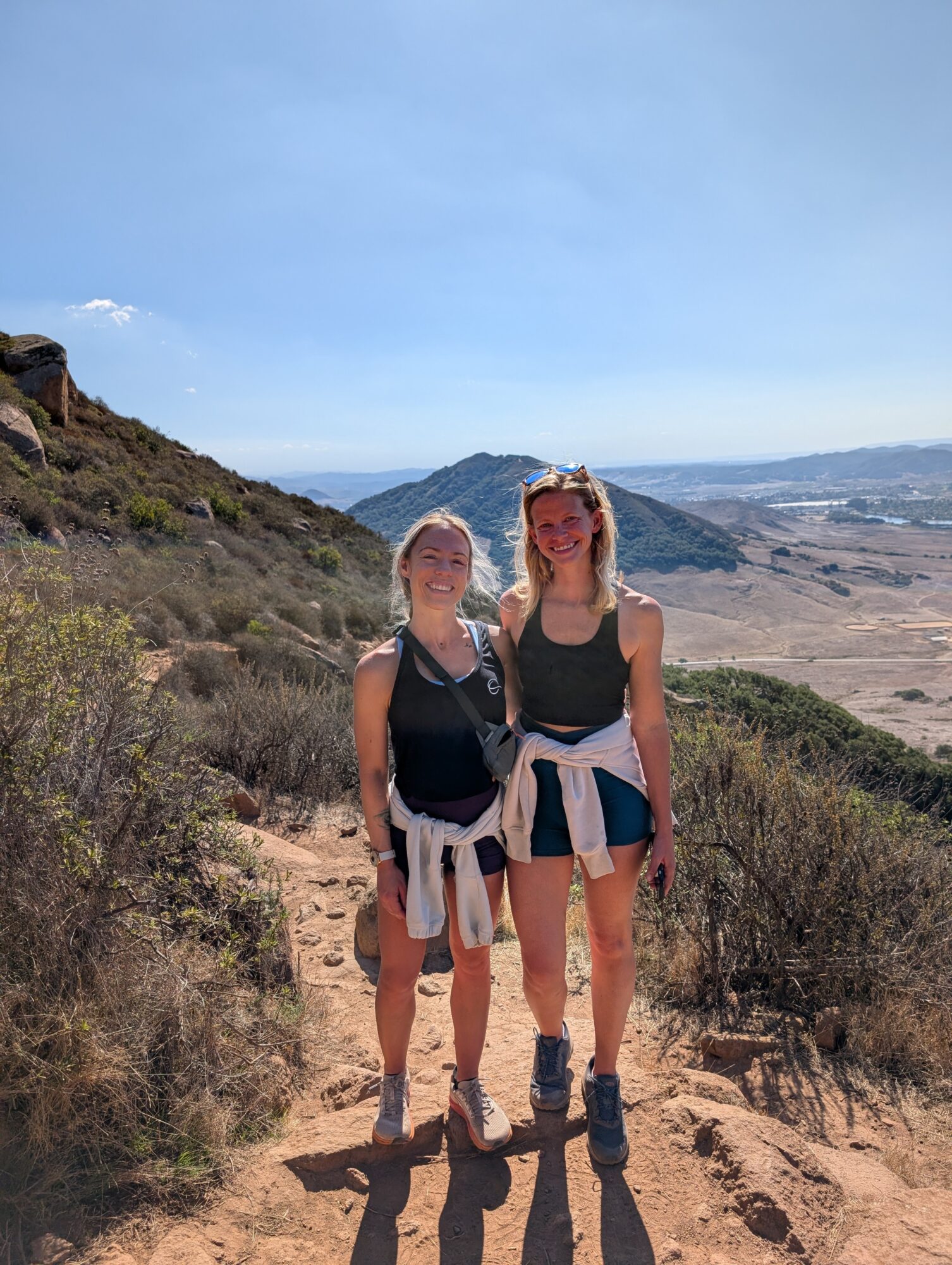 Two women stand on a dirt trail with hills and a distant landscape behind them, smiling at the camera.