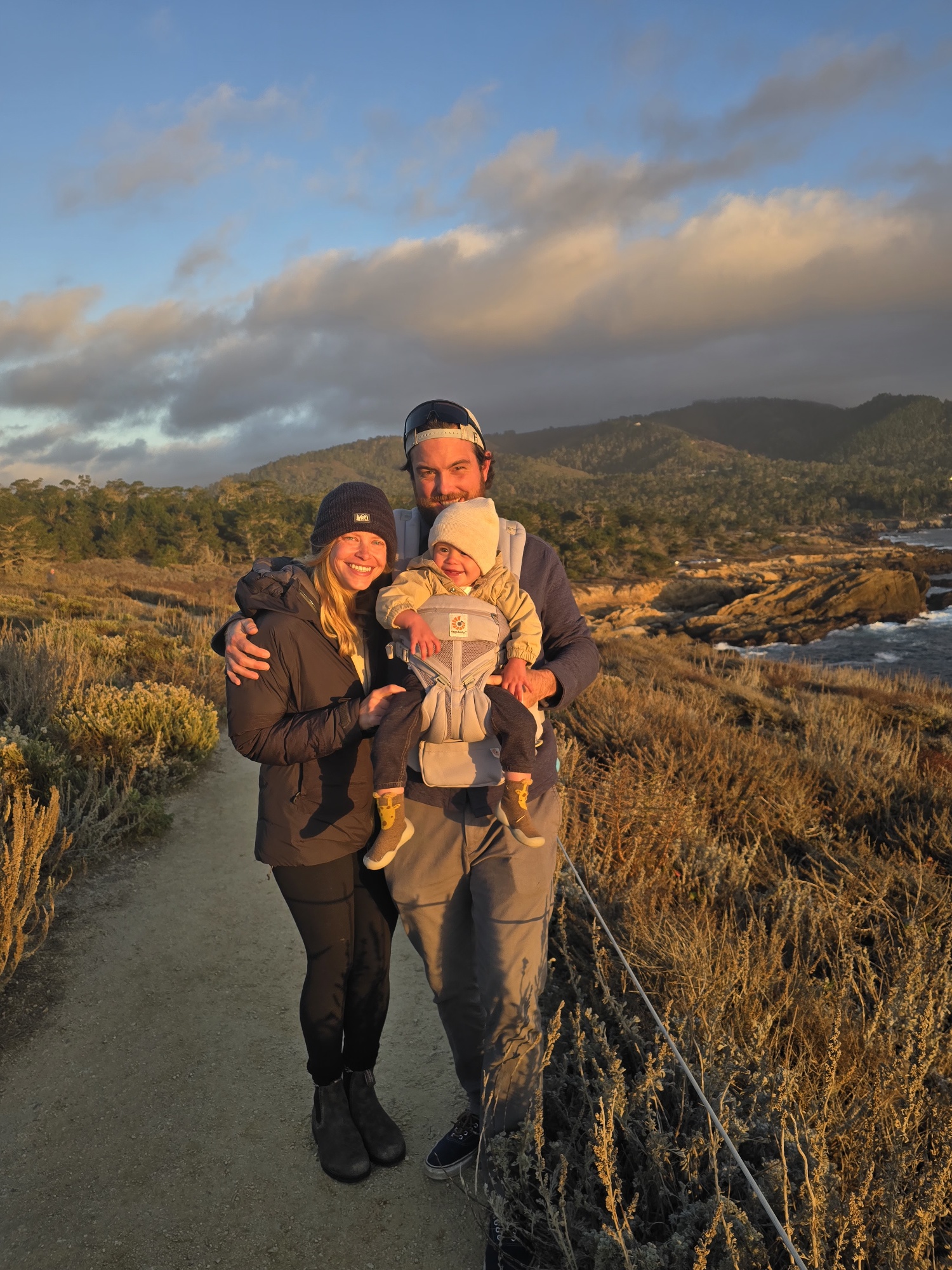 Family of three standing on a trail in a natural landscape with mountains and sky in the background.