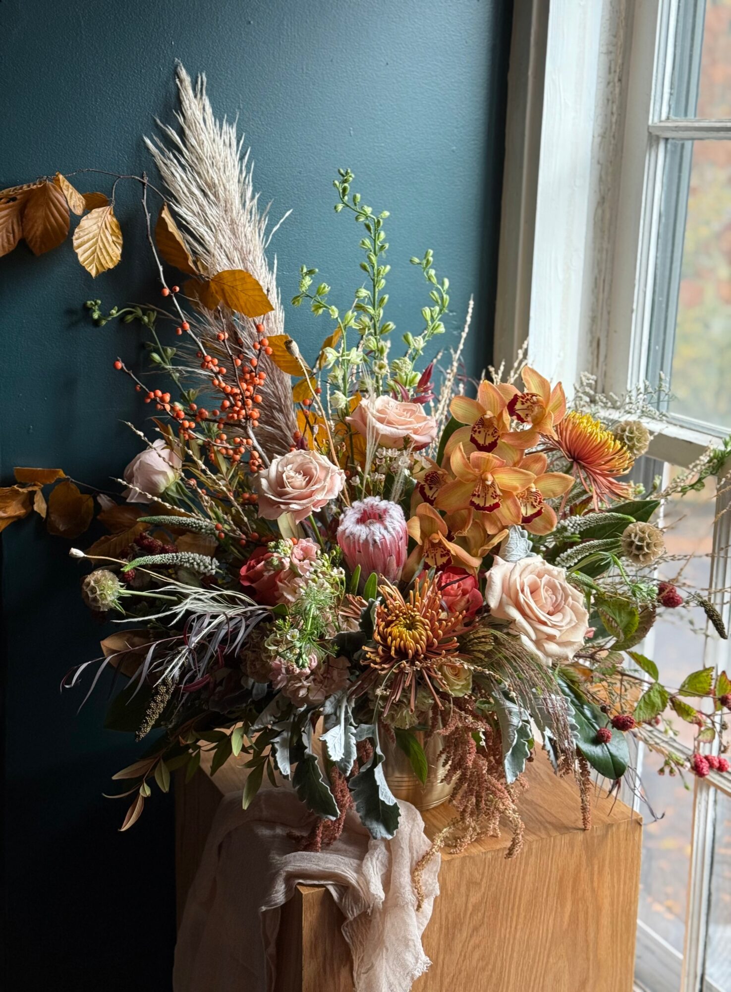 A floral arrangement with pink roses, dried leaves, and grasses on a wooden surface near a window.