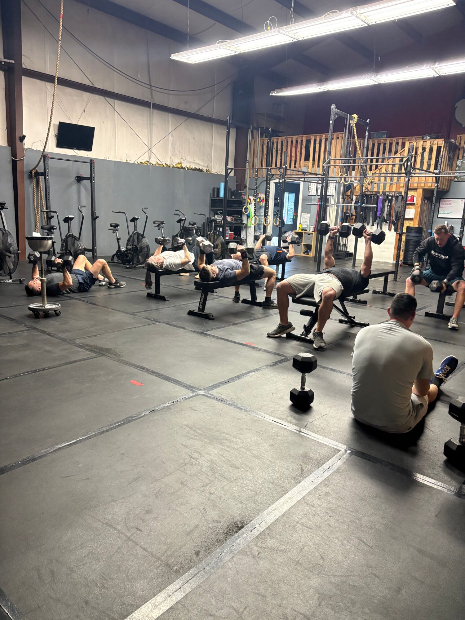 People working out with weightlifting equipment in a gym, some sitting and some standing, with gym machines and mirrors in the background.