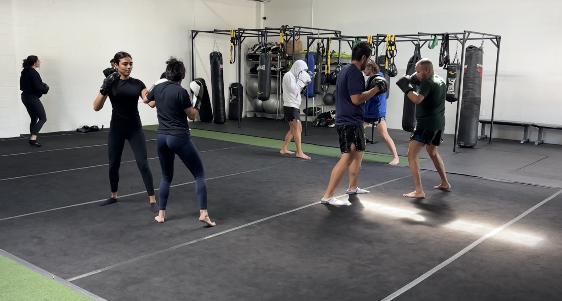 People practicing martial arts in a gym with training equipment, some sparring and others observing, on a black mat floor.