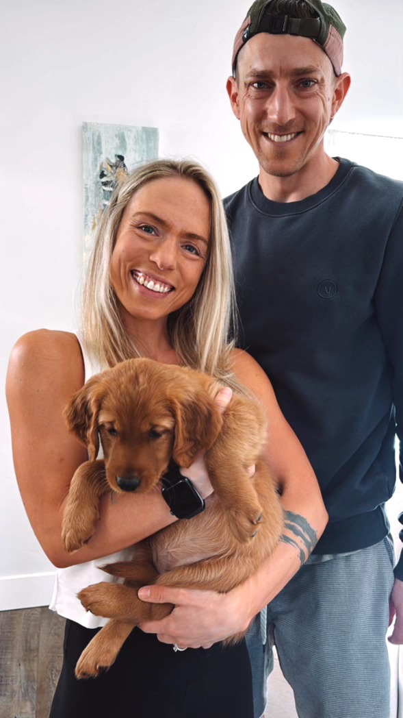 Smiling woman holding a puppy, with a smiling man standing beside them in a bright room.