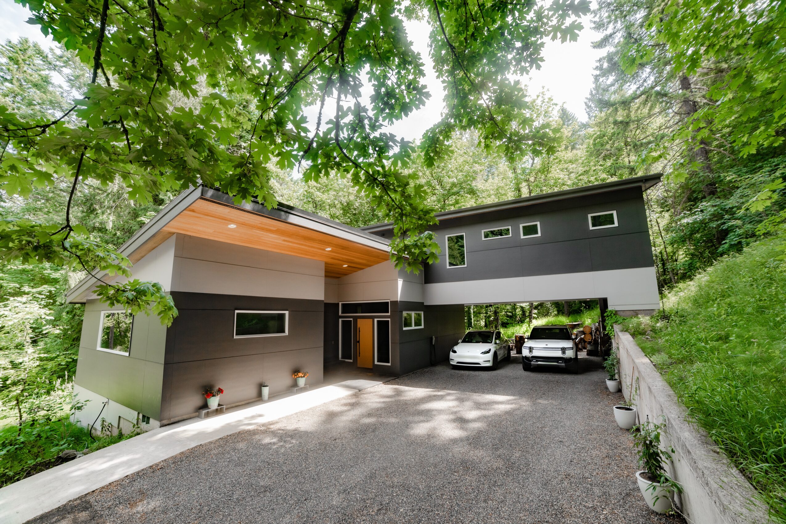Modern house with gray exterior, surrounded by green trees, with a driveway and parked cars, in a wooded area.