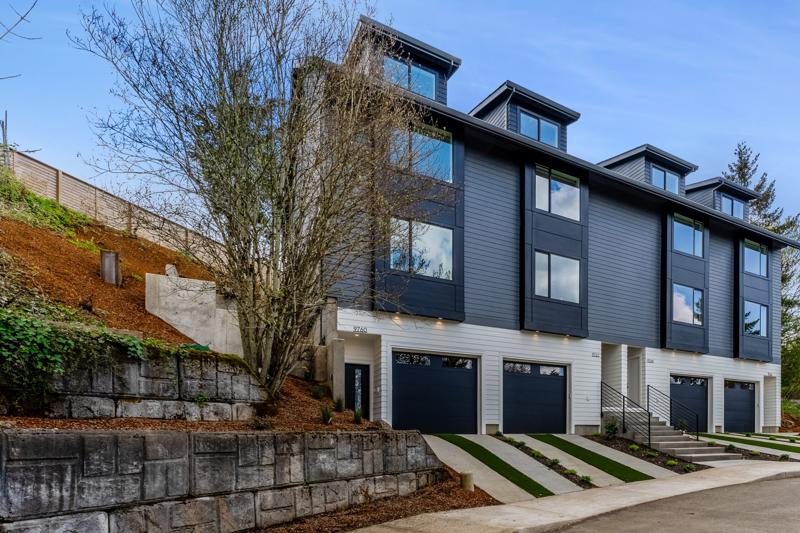 Multi-story modern building with garages, large windows, and a tree in front, against a blue sky.