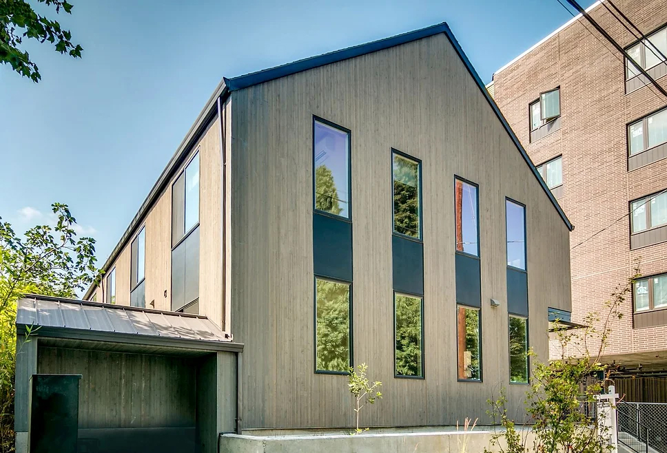 Modern building with tall, narrow windows and a sloped roof, surrounded by trees and neighboring structures.