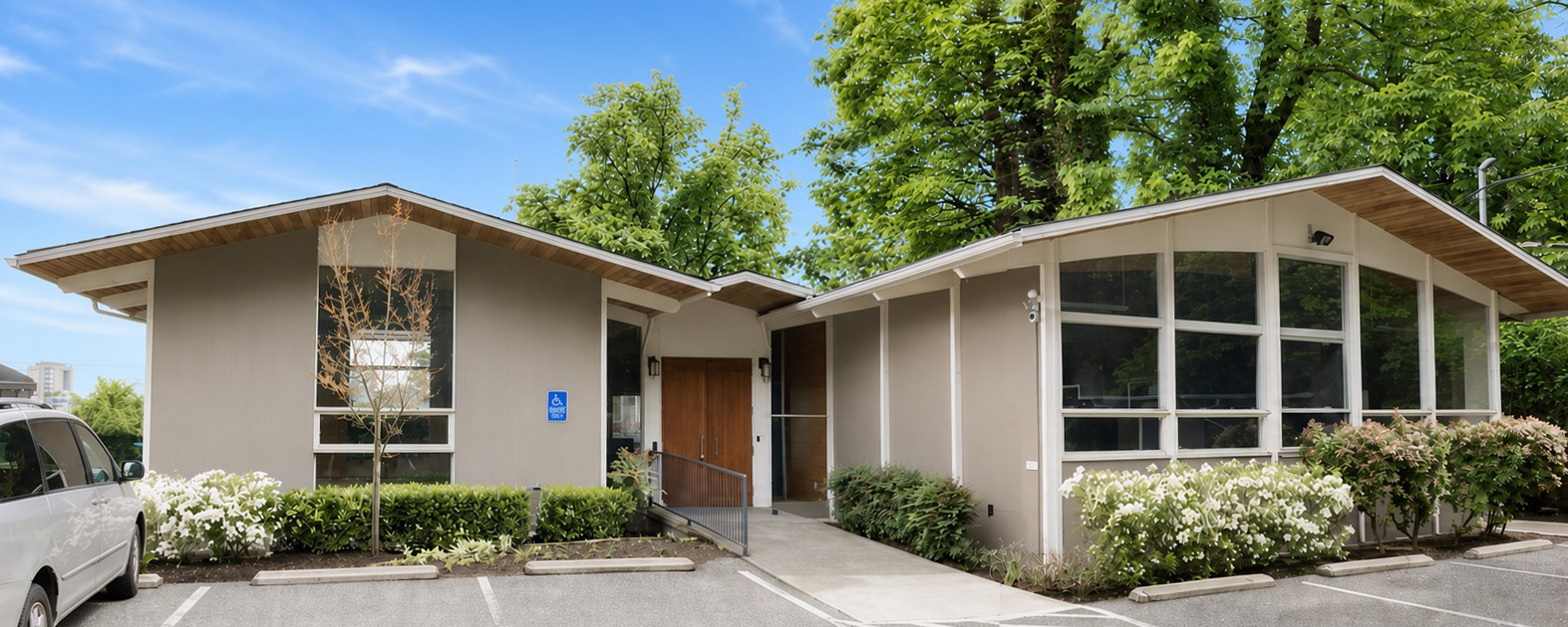 Single-story building with large windows, surrounded by greenery and bushes, under a blue sky.
