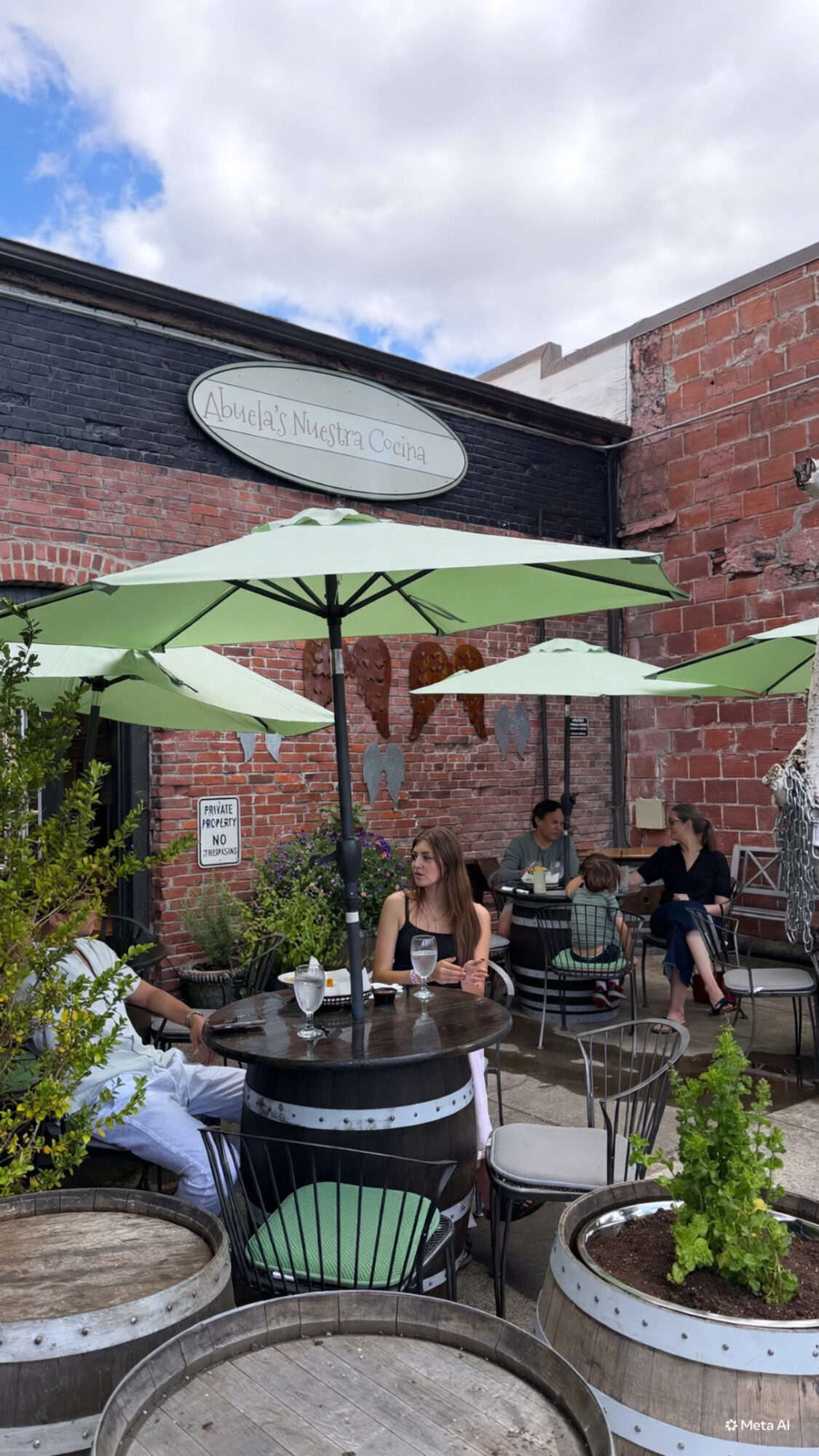 People sitting at outdoor tables with umbrellas in a courtyard area, brick walls and a sign above.
