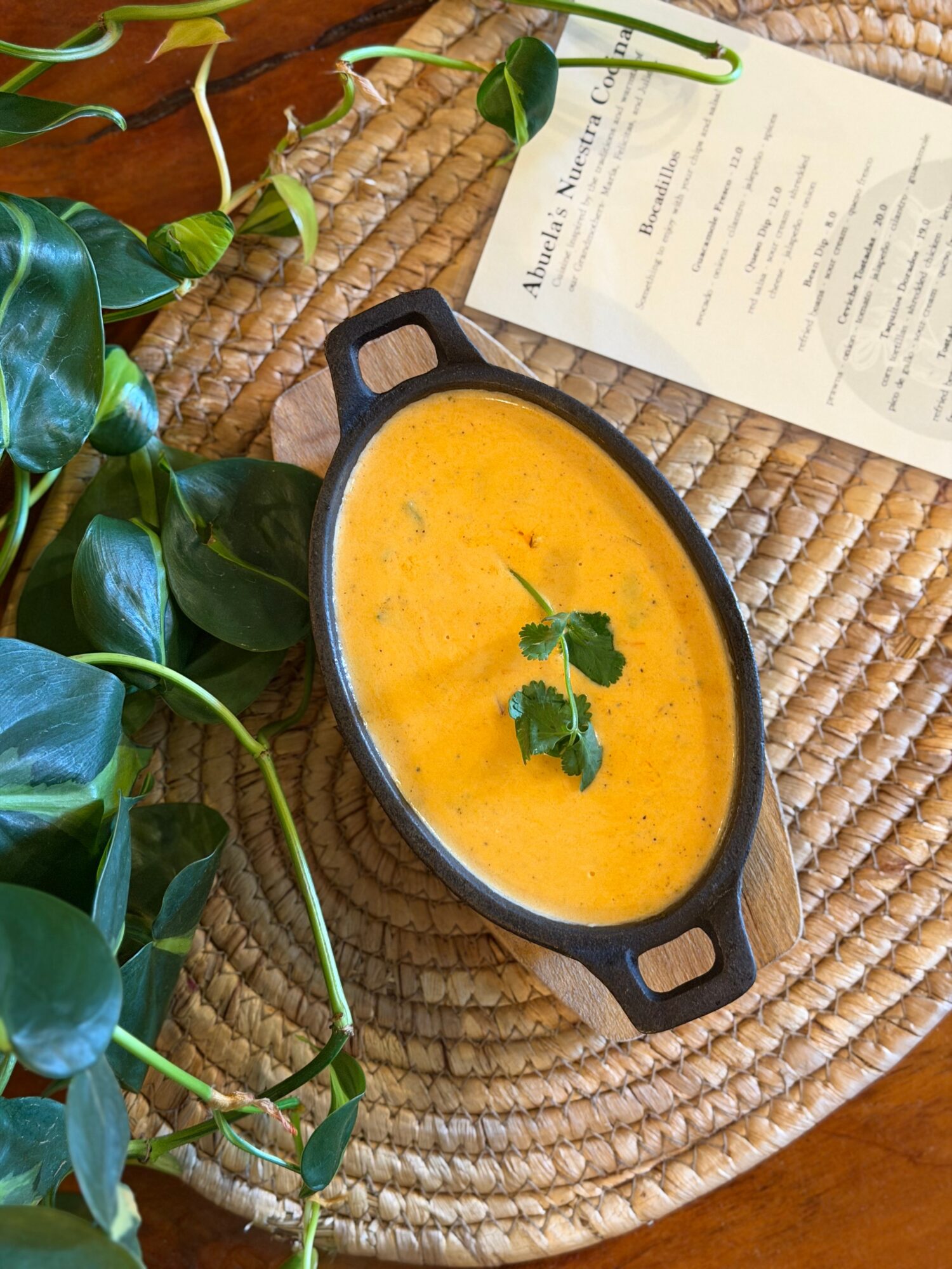 Yellow soup in a black bowl with a green leaf garnish, placed on a woven mat with a plant and paper nearby.